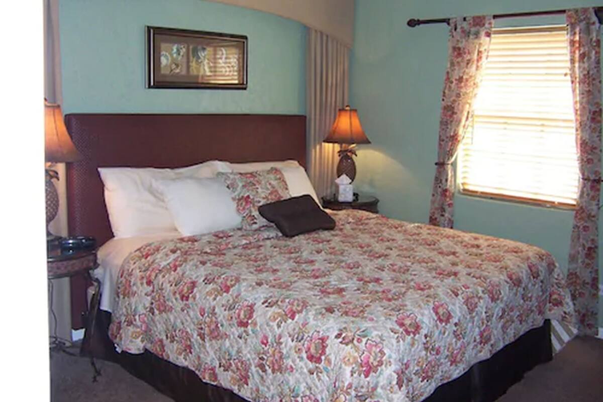 A bedroom with a bed, a nightstand, a lamp, a window with curtains, and a framed picture on the wall.