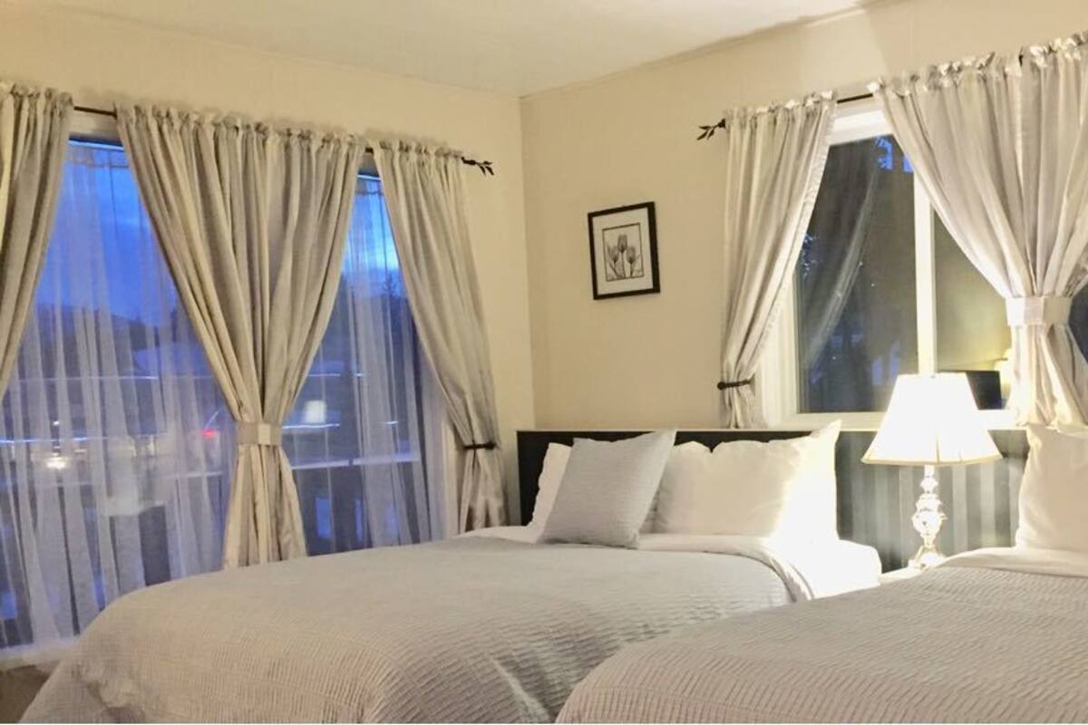 A hotel room with two beds, a nightstand with a lamp, and a window with curtains.