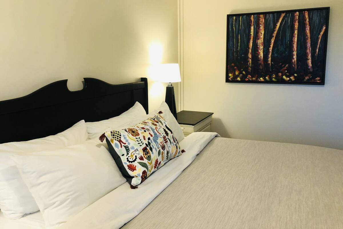 A neatly made bed with white linens and a colorful floral pillow, a black headboard, a bedside table with a lamp, and a painting of trees on the wall.
