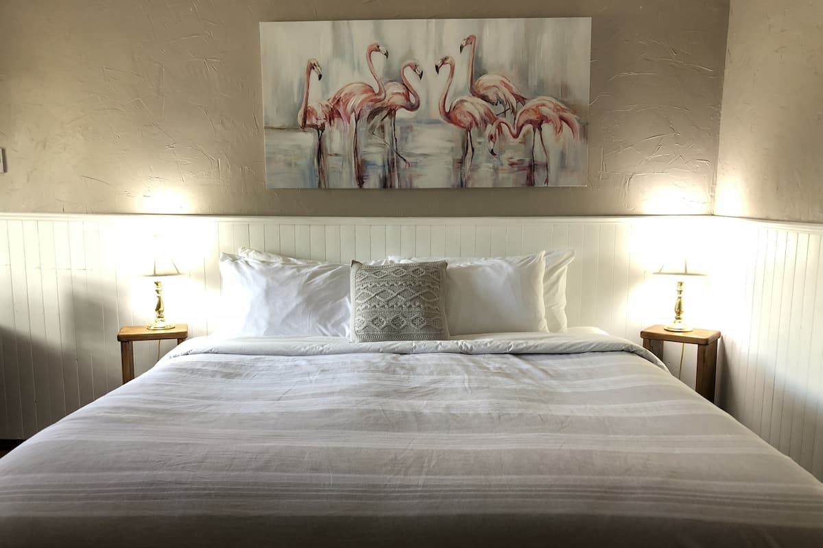 A neatly made bed with white linens, two bedside lamps, and a painting of flamingos on the wall.