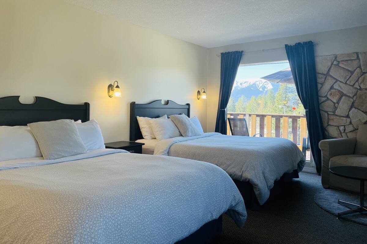 A hotel room with two beds, a stone accent wall, a balcony with a view of mountains, and a chair.