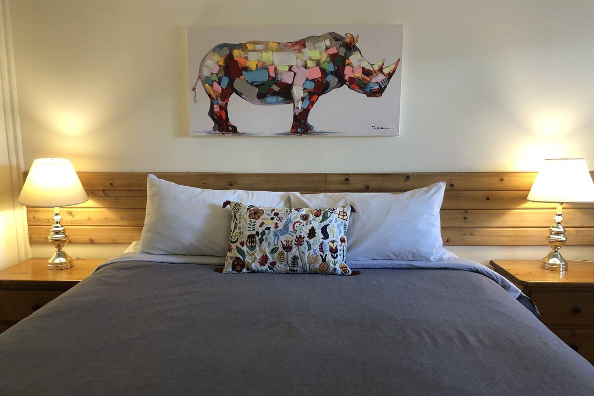 A bed with a grey bedspread, a colorful floral pillow, and a rhinoceros artwork on the wall.