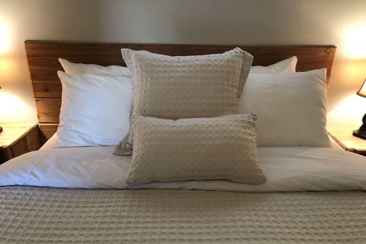 A neatly made bed with a textured white mattress and two pillows, a wooden headboard, and a nightstand with a lamp.