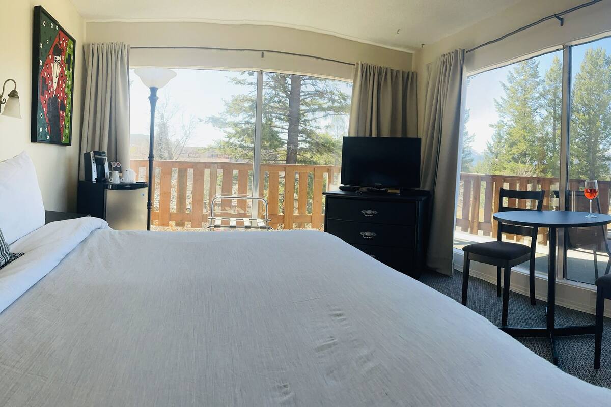 A hotel room with a bed, a TV, a desk with a chair, and a balcony with a view of trees and mountains.