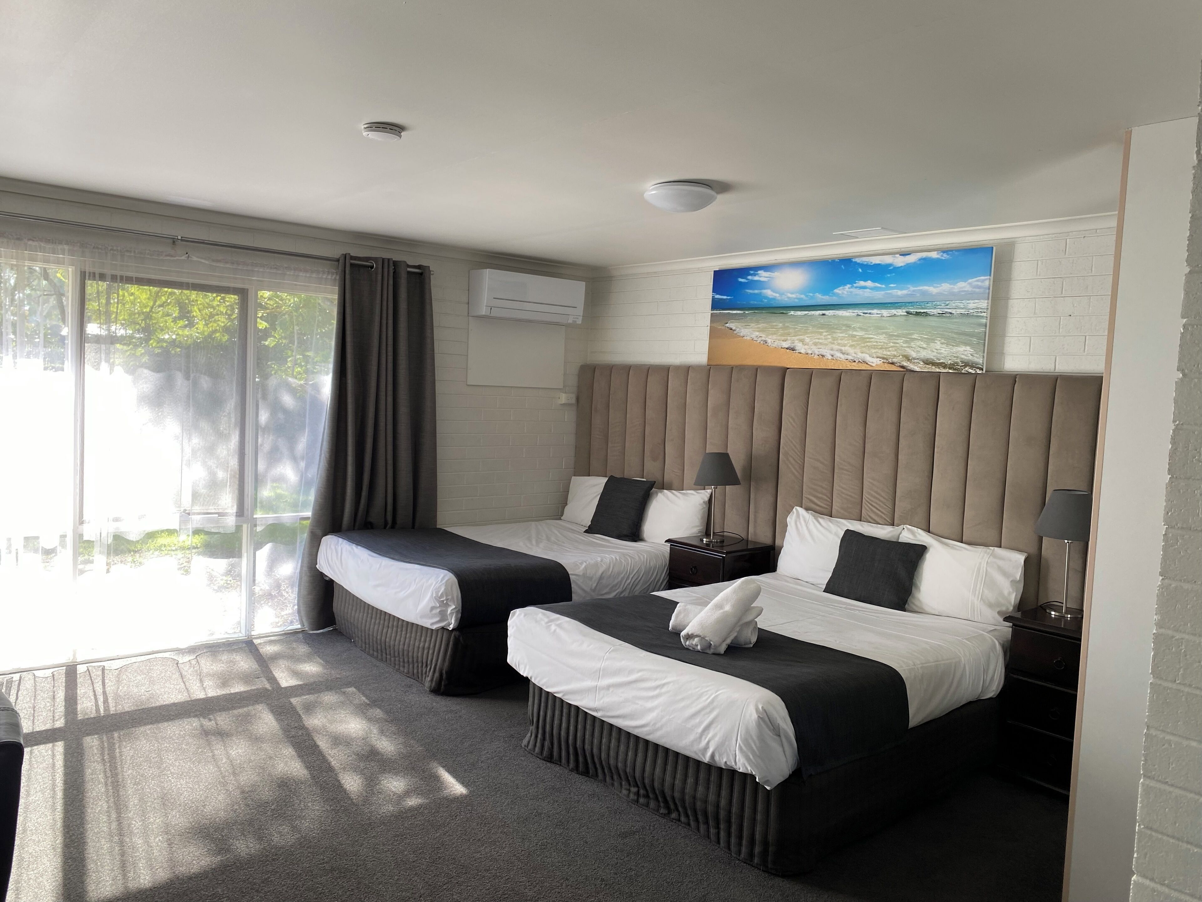 Een hotelkamer met twee bedden, een groot raam met gordijnen, een schilderij van een strand en een airconditioner.