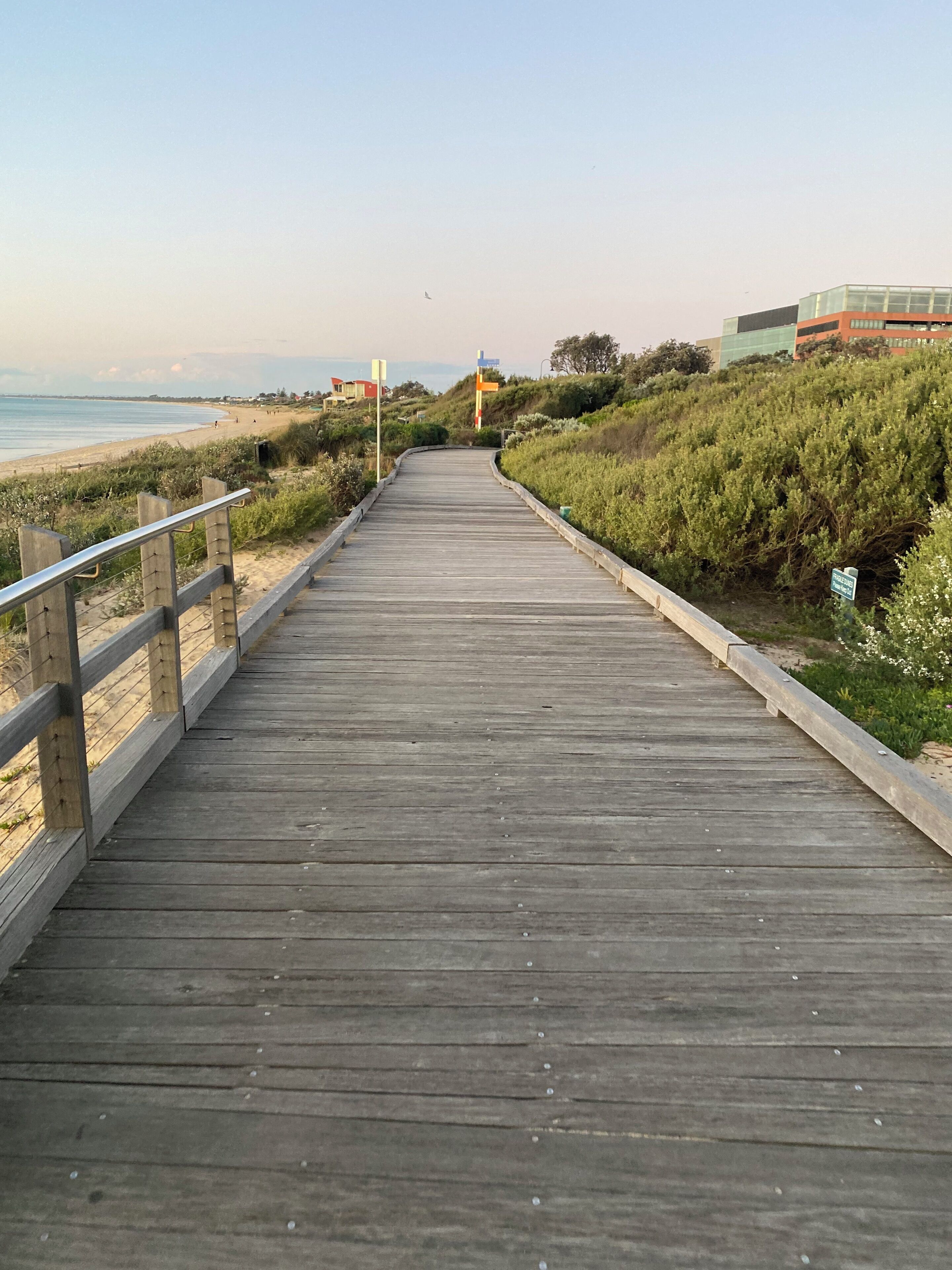 Una pasarela de madera que conduce a una playa, con una colina cubierta de hierba a un lado y un edificio al fondo.