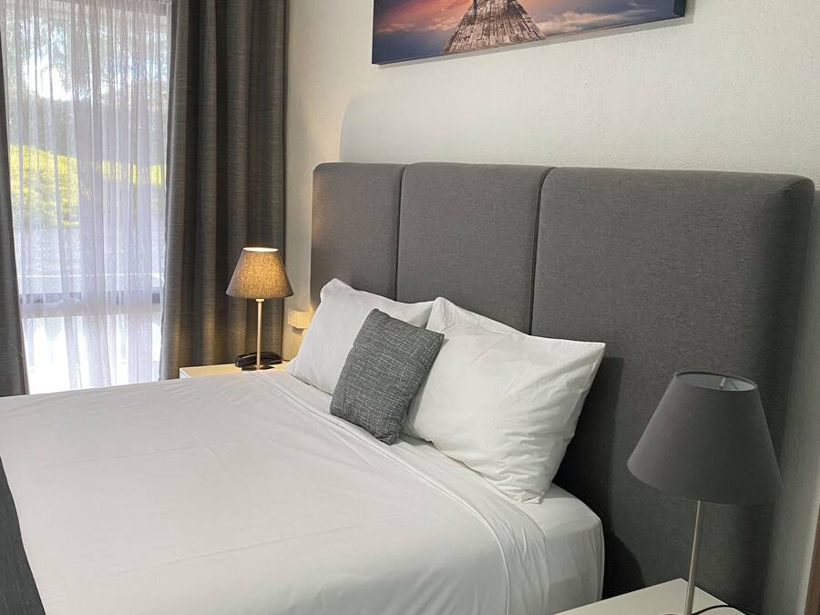 Una cama bien hecha con sábanas blancas y un cabecero gris, una lámpara de noche y un cuadro de una puesta de sol en la pared.