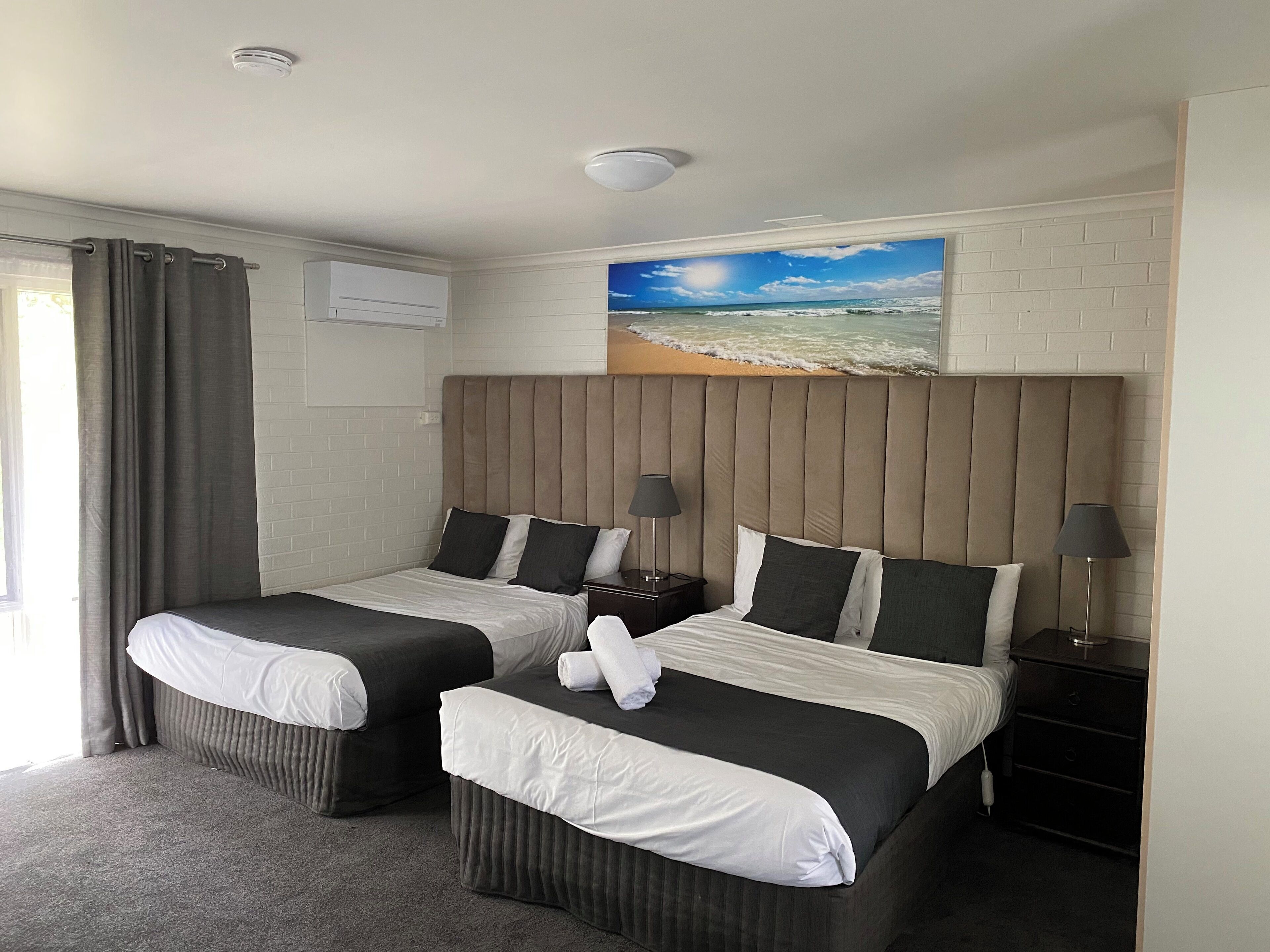 A hotel room with two beds, a large wall-mounted painting of a beach, and a window with curtains.