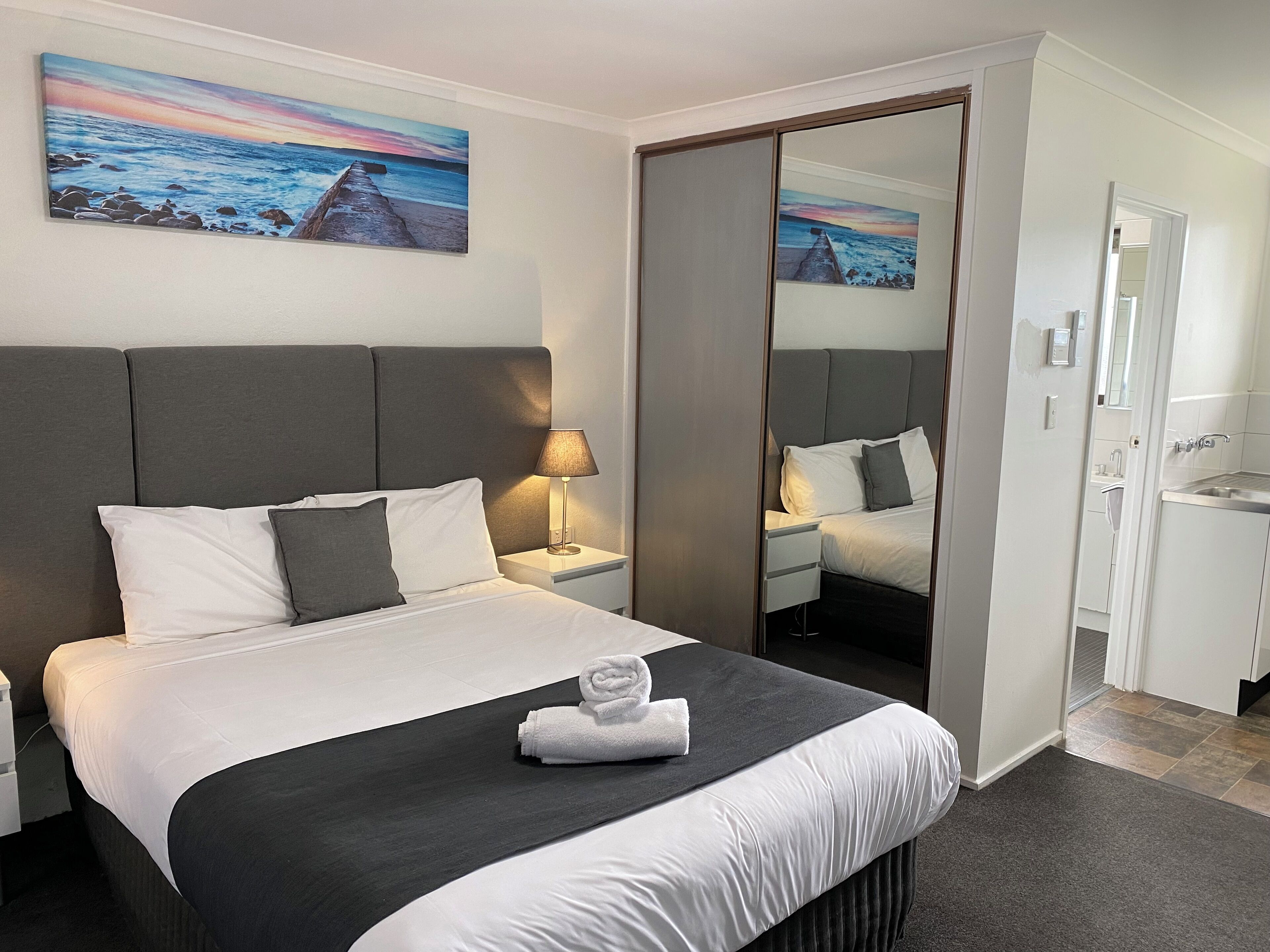 A modern hotel room with a large bed, a bedside table with a lamp, a mirror, and a painting of a beach scene.