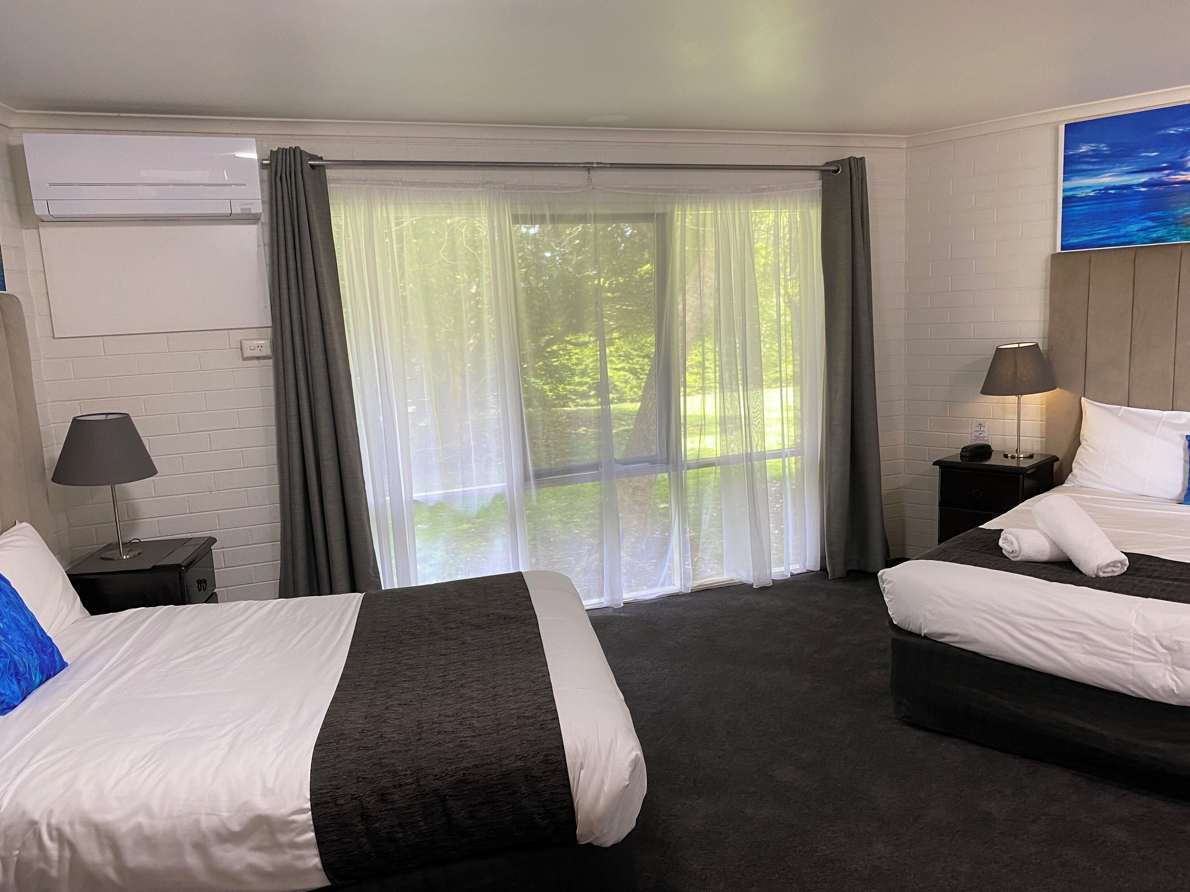 Une chambre d’hôtel avec deux lits, une grande fenêtre avec des rideaux légers, un climatiseur fixé au mur, une table de chevet avec une lampe et un tableau accroché au mur.