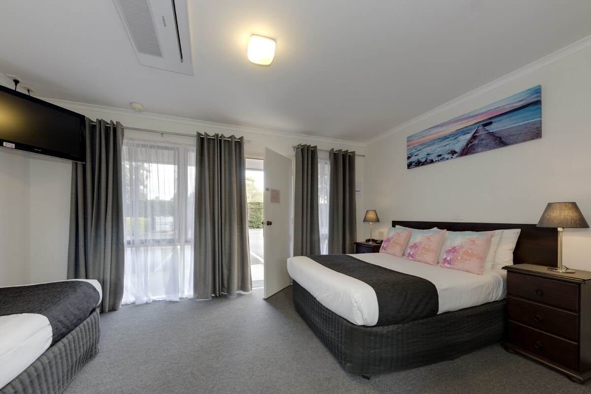 A hotel room with a bed, a bedside table with a lamp, a painting on the wall, and a view of the outdoors through the window.
