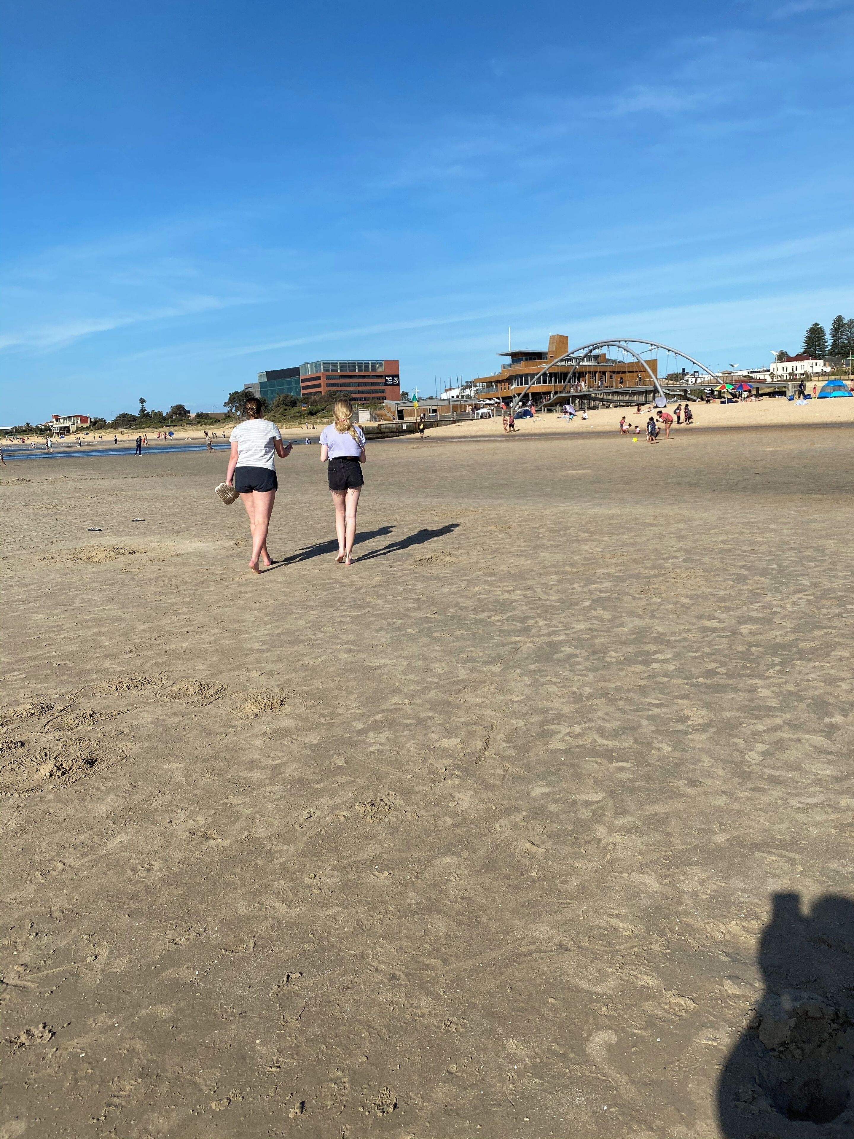 A sandy beach with a few people walking. There are buildings and a large structure in the background.