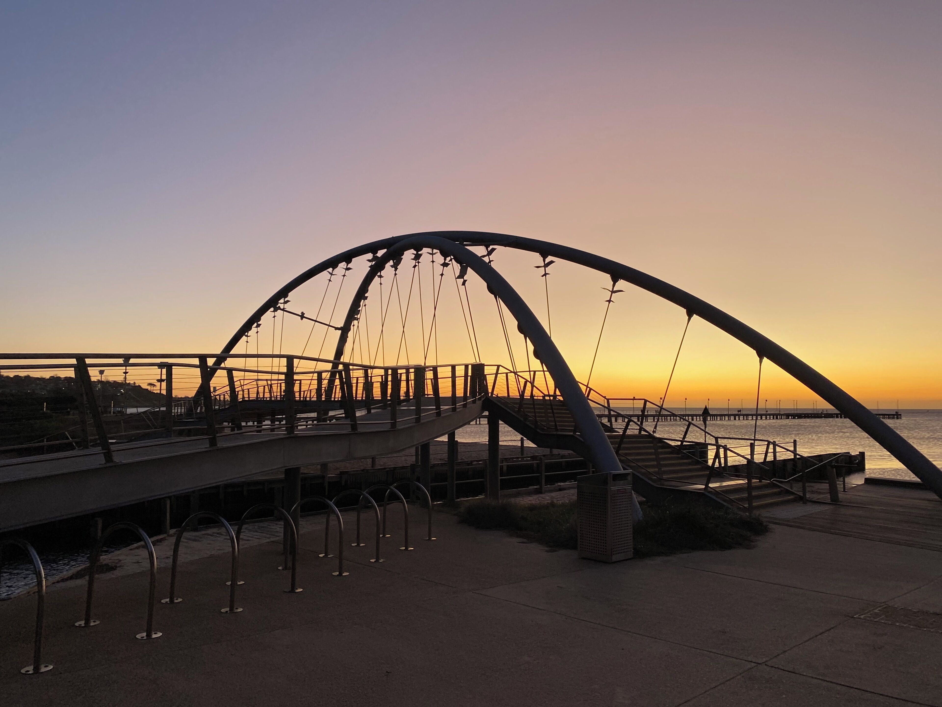Un puente peatonal con un arco metálico curvo, que une dos extensiones de tierra sobre una masa de agua.