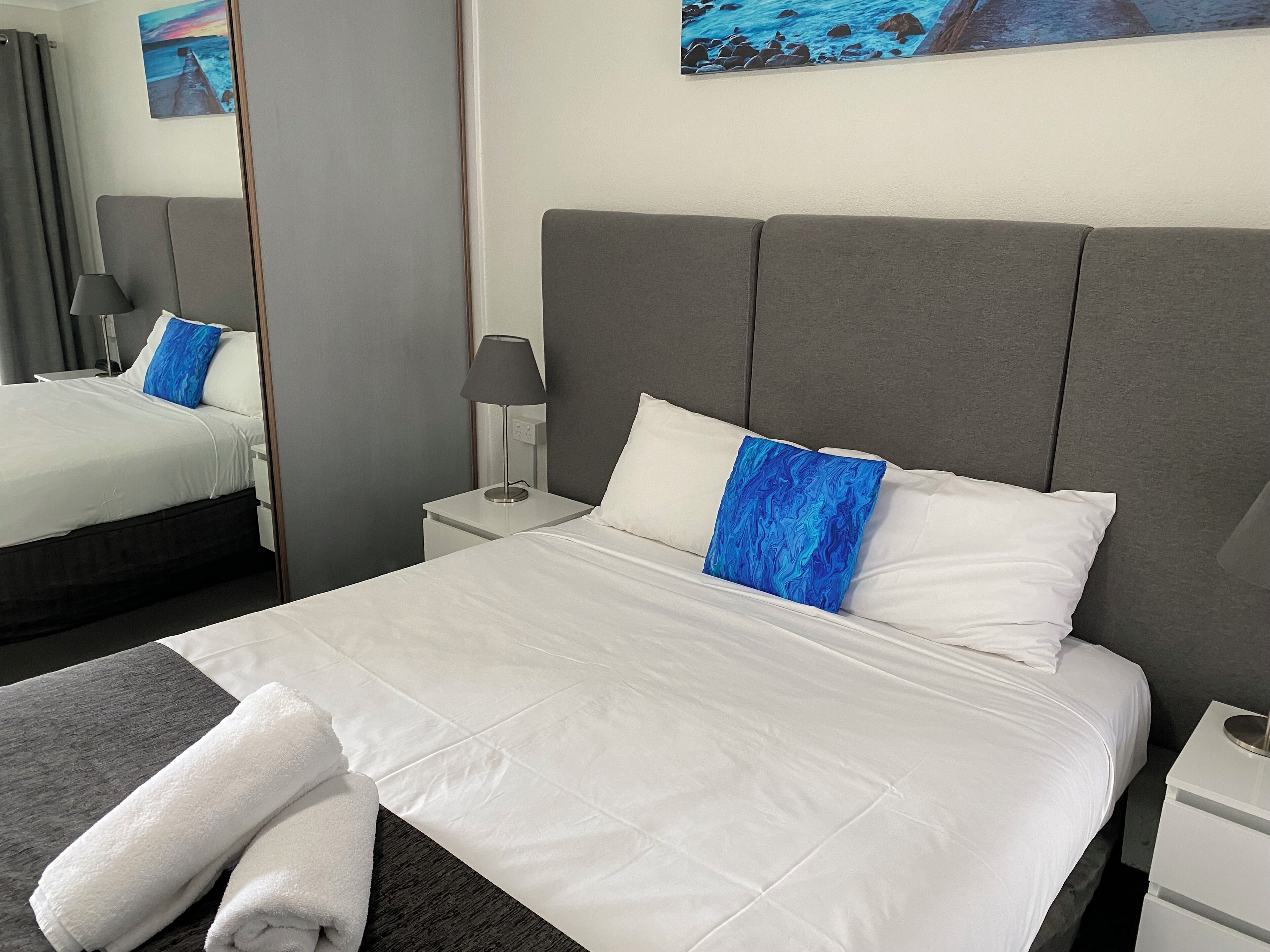 A neatly made bed with white linens and a blue pillow, a bedside lamp, and a small white nightstand.