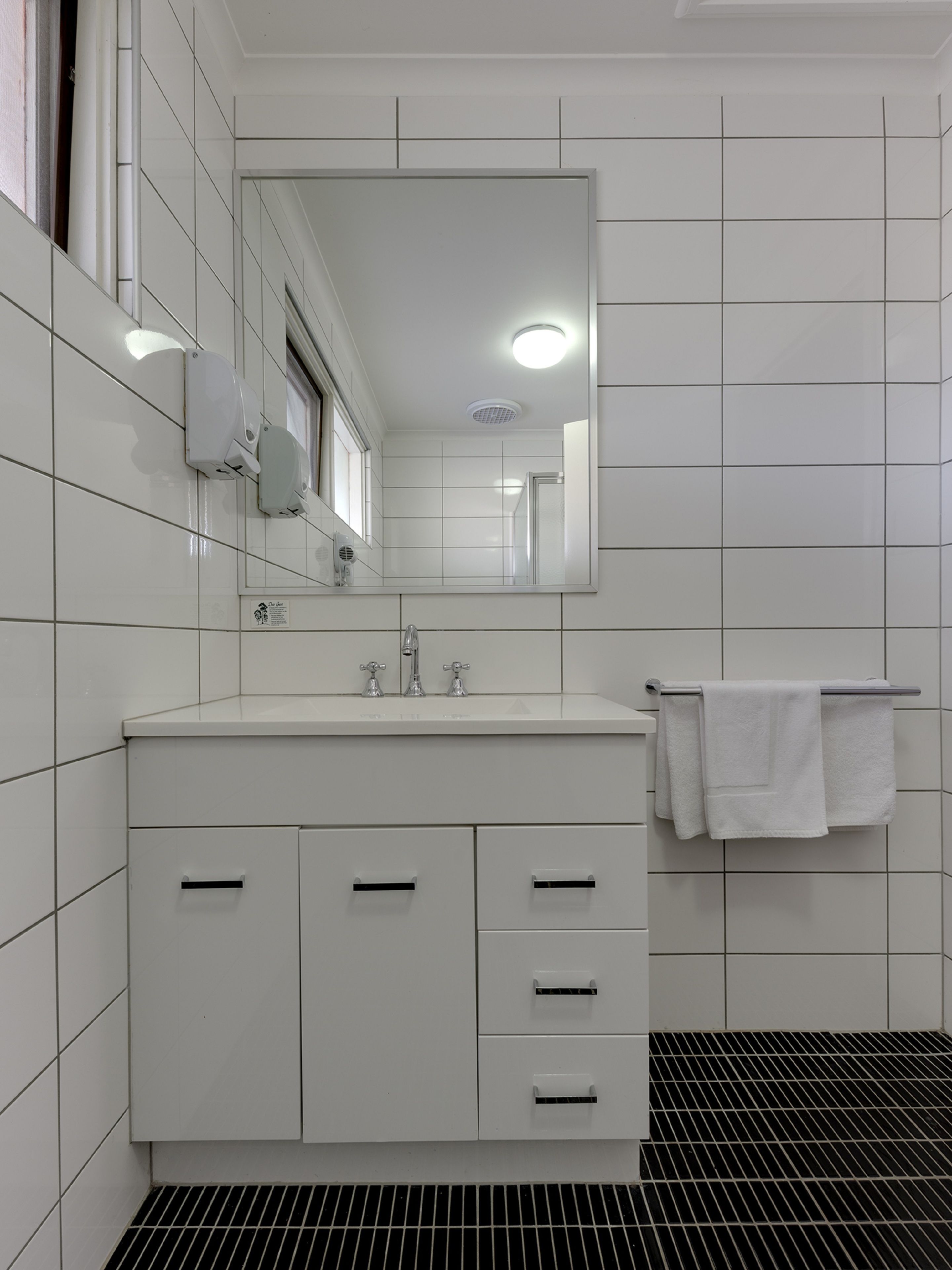 Ein modernes Badezimmer mit weißen Fliesen, einem großen Spiegel, einem Waschbecken mit zwei Wasserhähnen und einem Handtuchhalter mit einem weißen Handtuch.