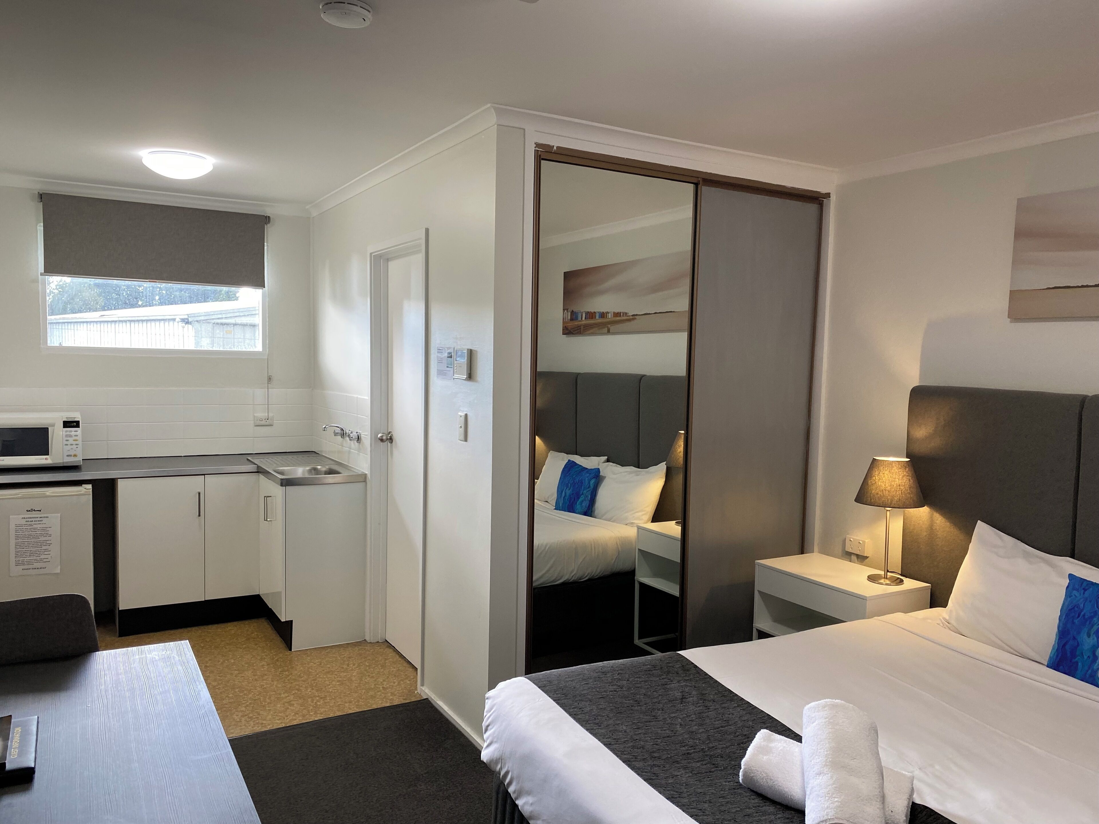 A hotel room with a bed, bedside table, lamp, and a kitchenette with a microwave.