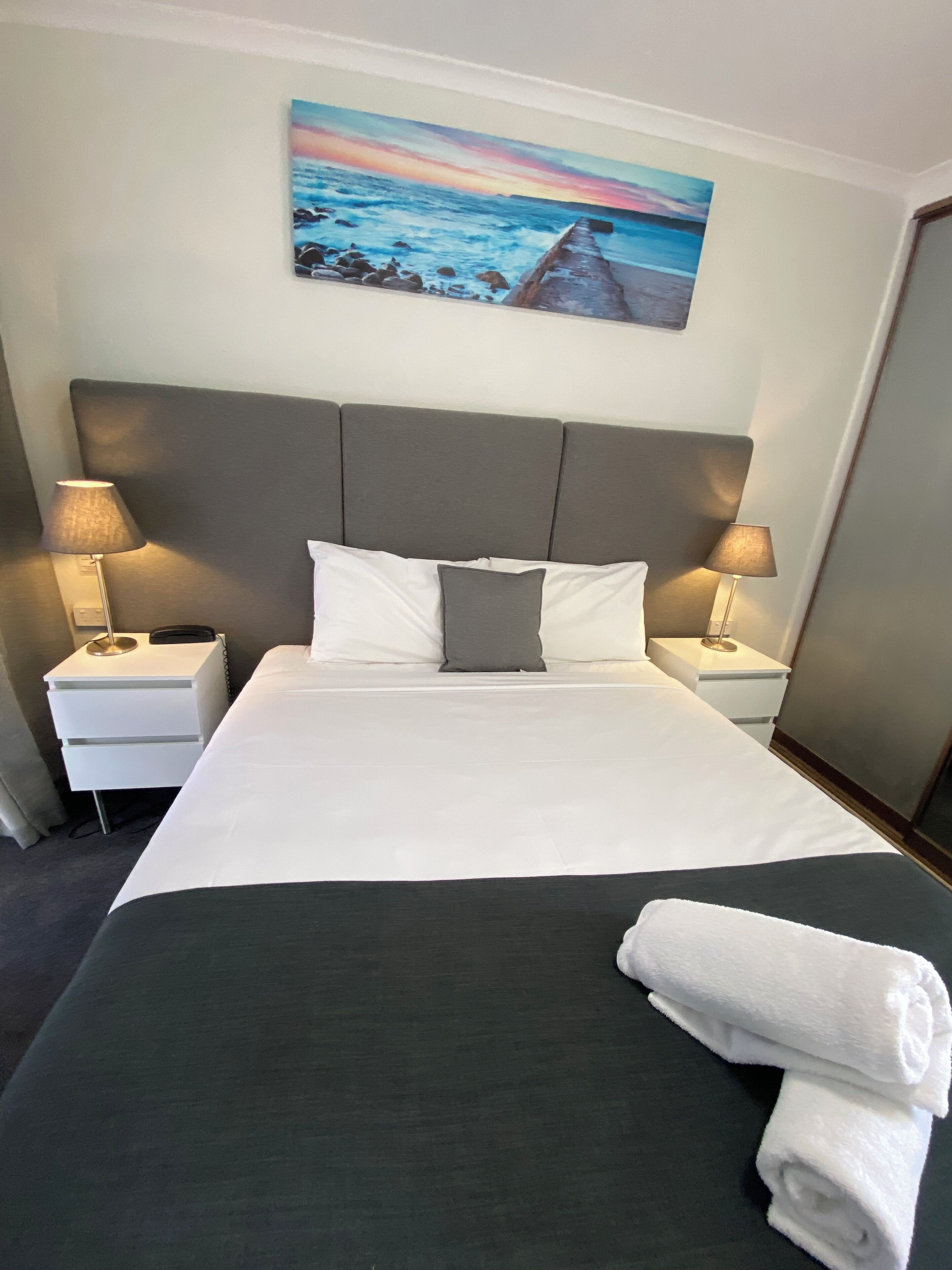 A hotel room with a large bed, two bedside tables, a headboard, and a painting of a seascape.