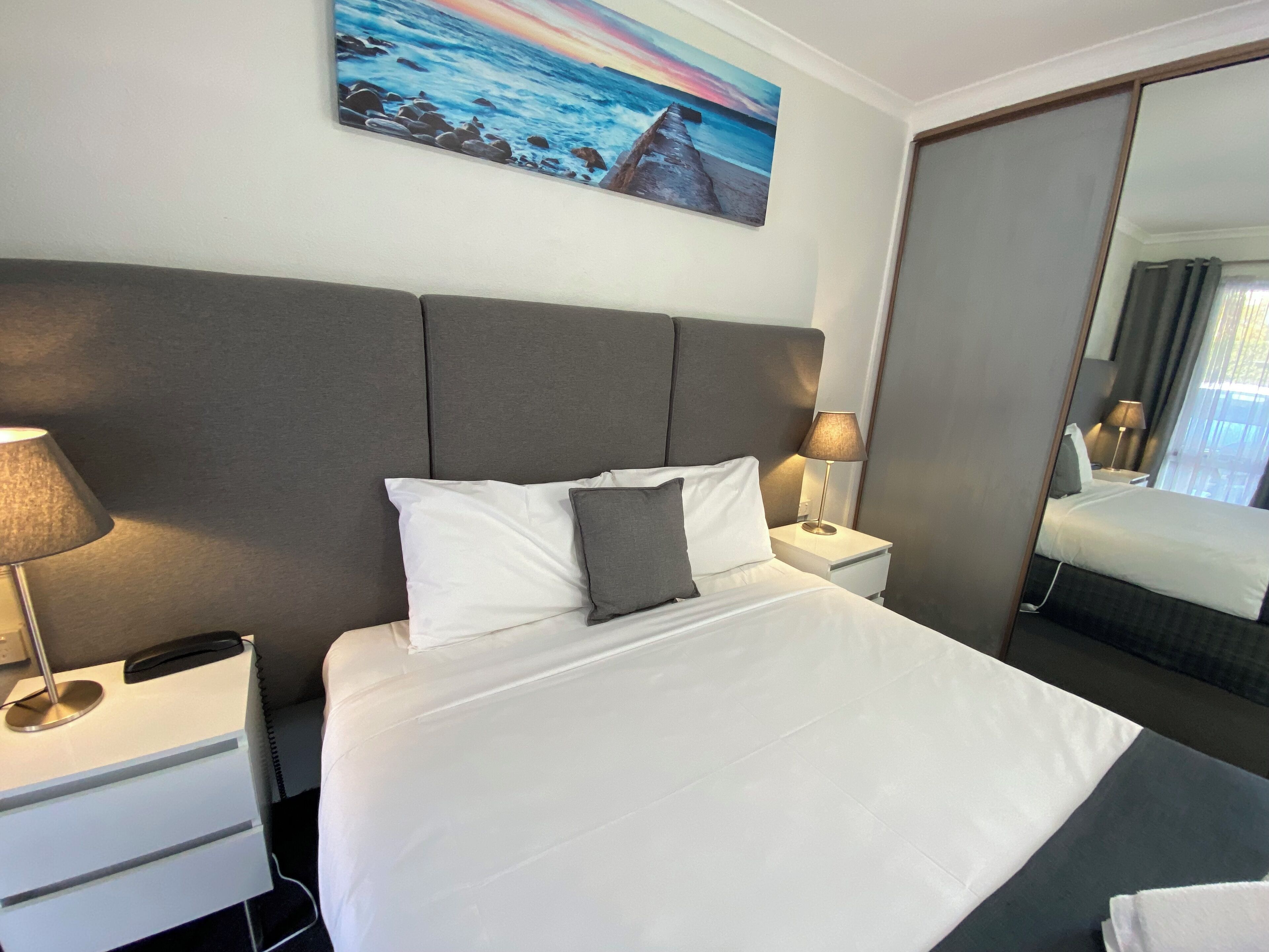 Ein Hotelzimmer mit einem großen Bett, einem Nachttisch mit Lampe, einem Telefon und einem Strandbild an der Wand.