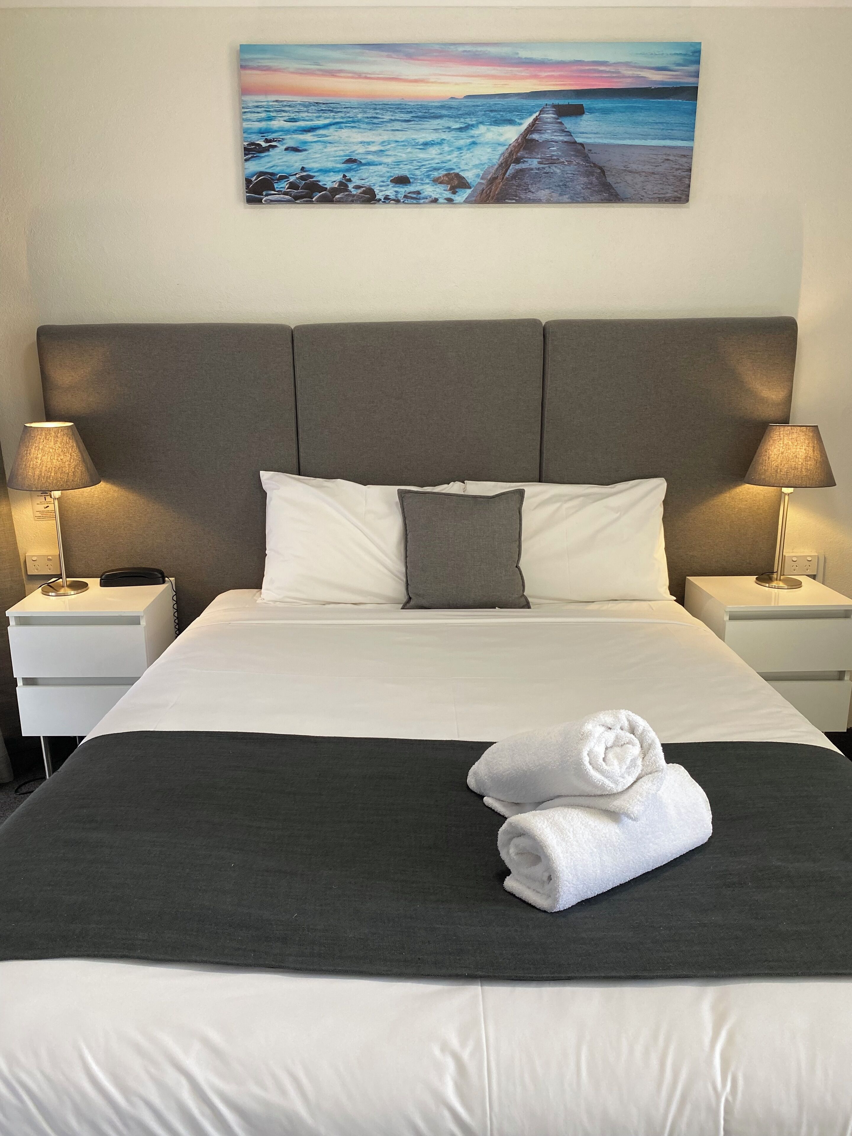 A neatly made bed with white and gray linens, two bedside lamps, and a painting of a coastal sunset above the bed.