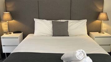 A neatly made bed with white and gray linens, two bedside lamps, and a painting of a coastal sunset above the bed.