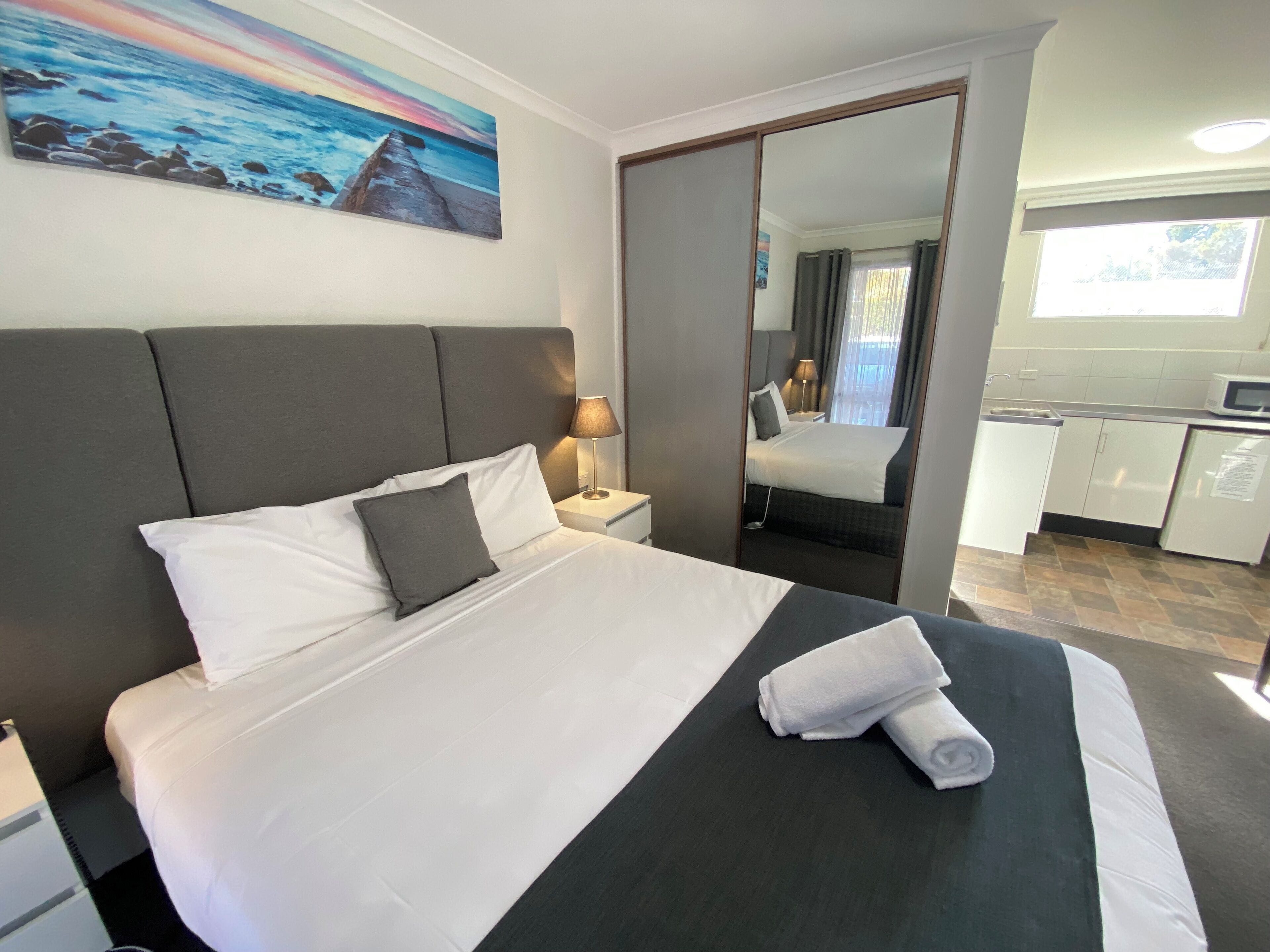 A hotel room with a large bed, a nightstand, a mirror, a view of the kitchen, and a painting on the wall.