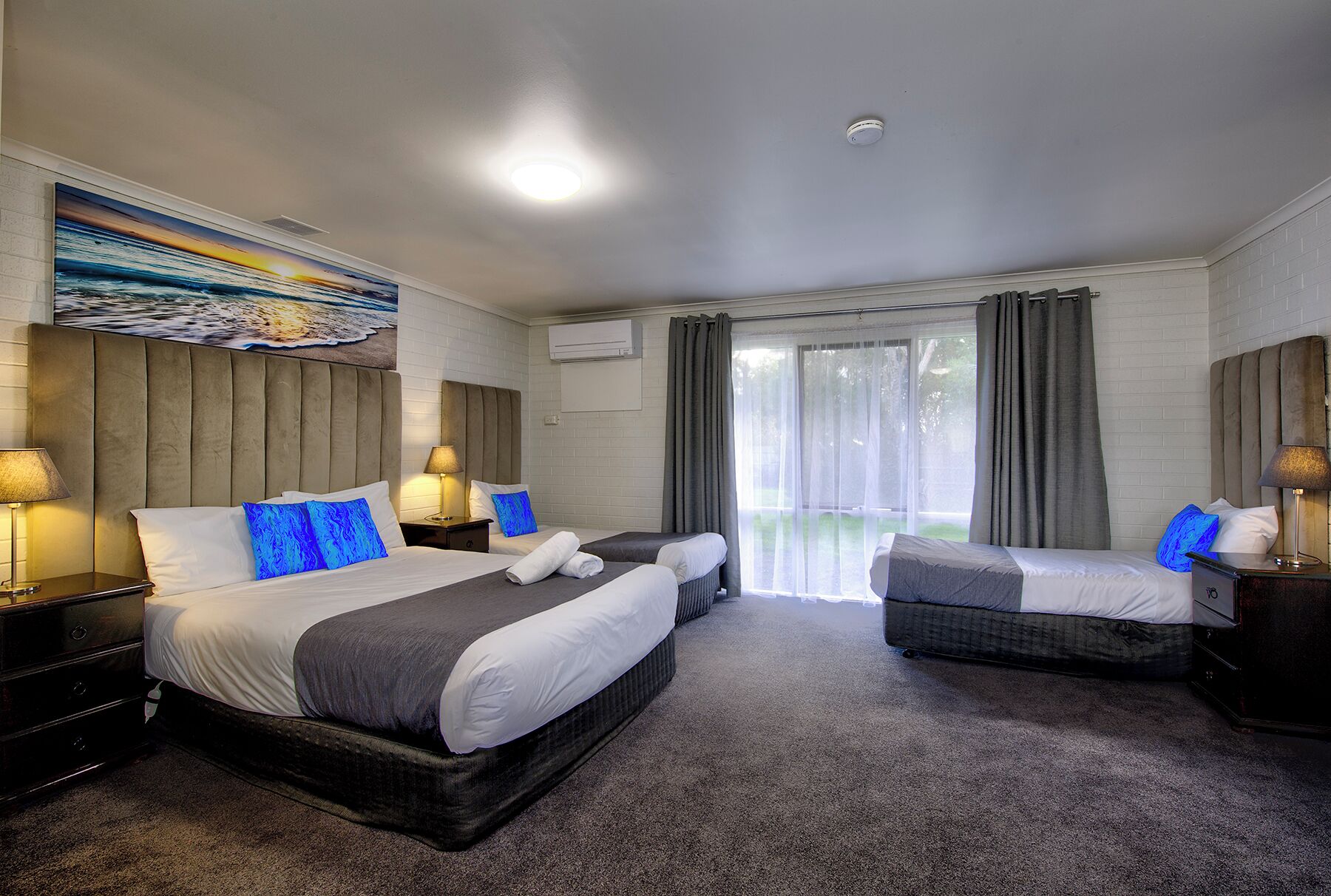 Habitación de hotel con dos camas, un ventanal con cortinas, un cuadro en la pared y una lámpara en el techo.