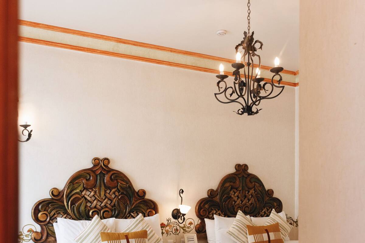 A hotel room with two beds, ornate wooden headboards, a chandelier, and a tiled floor.