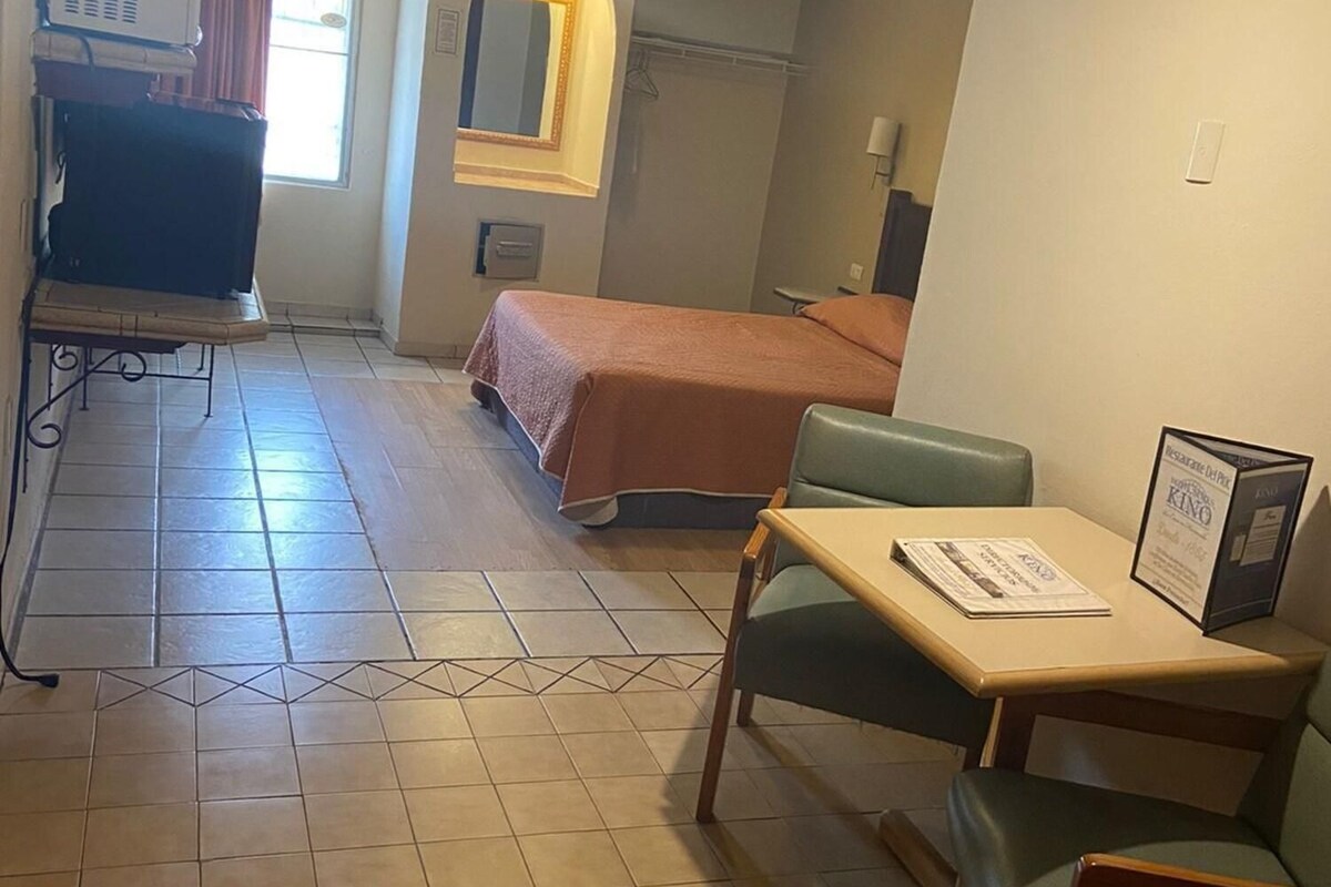Habitación de hotel con cama, escritorio con silla, ventilador de techo, televisión y ventana con cortinas.