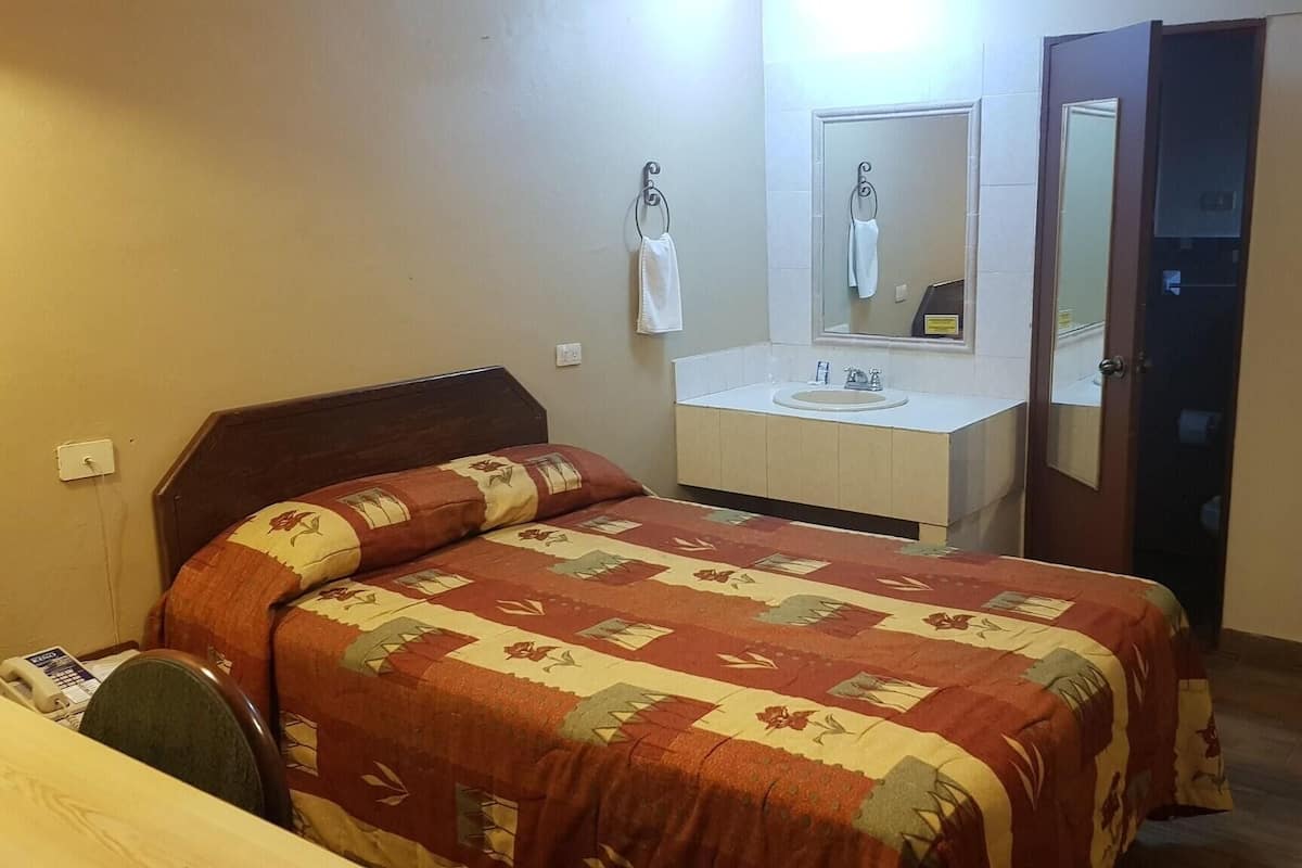 Una habitación de hotel con una cama, un baño con lavamanos y espejo, y una puerta.