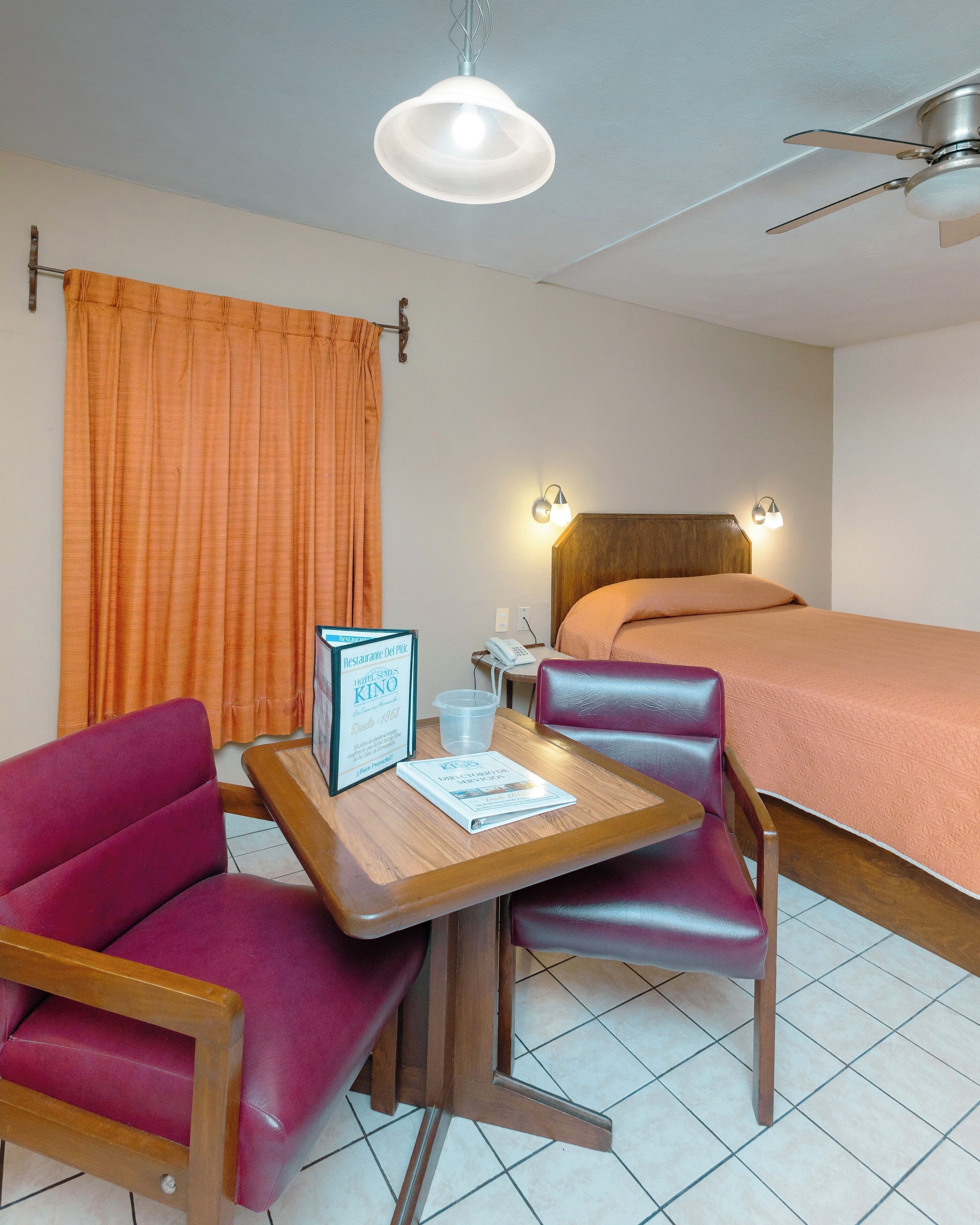 A hotel room with a bed, a desk with two chairs, a ceiling fan, and a window with curtains.
