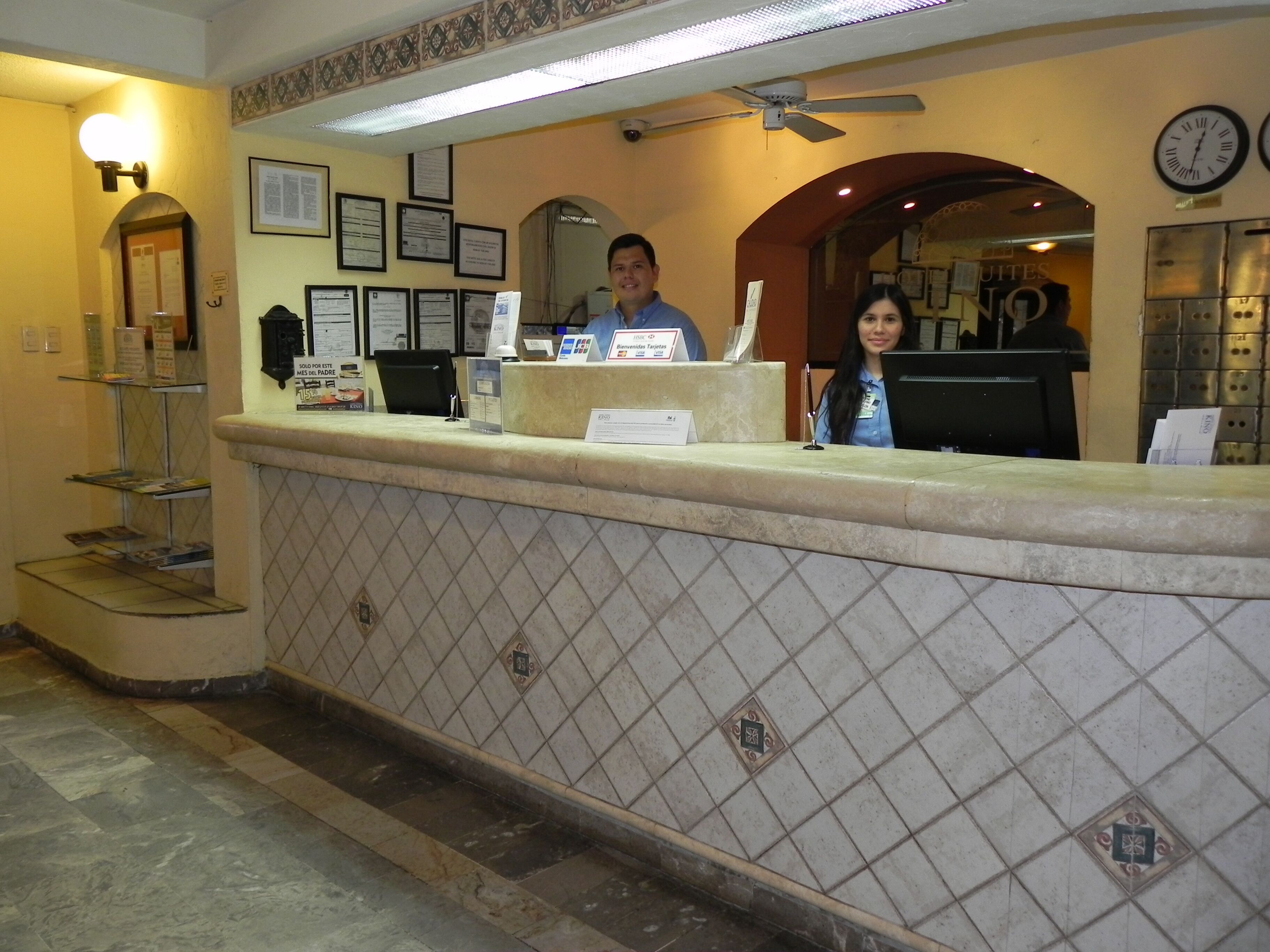 Zona de recepción con pared de azulejos, mostrador de recepción, certificados enmarcados y un reloj.