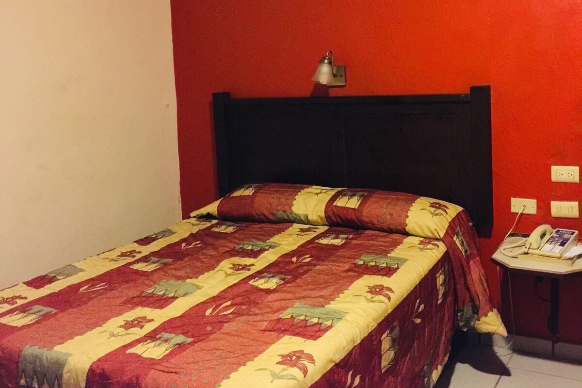 Un dormitorio con una pared roja, una cama con colcha estampada, una mesita de noche con teléfono y una lámpara de pared.