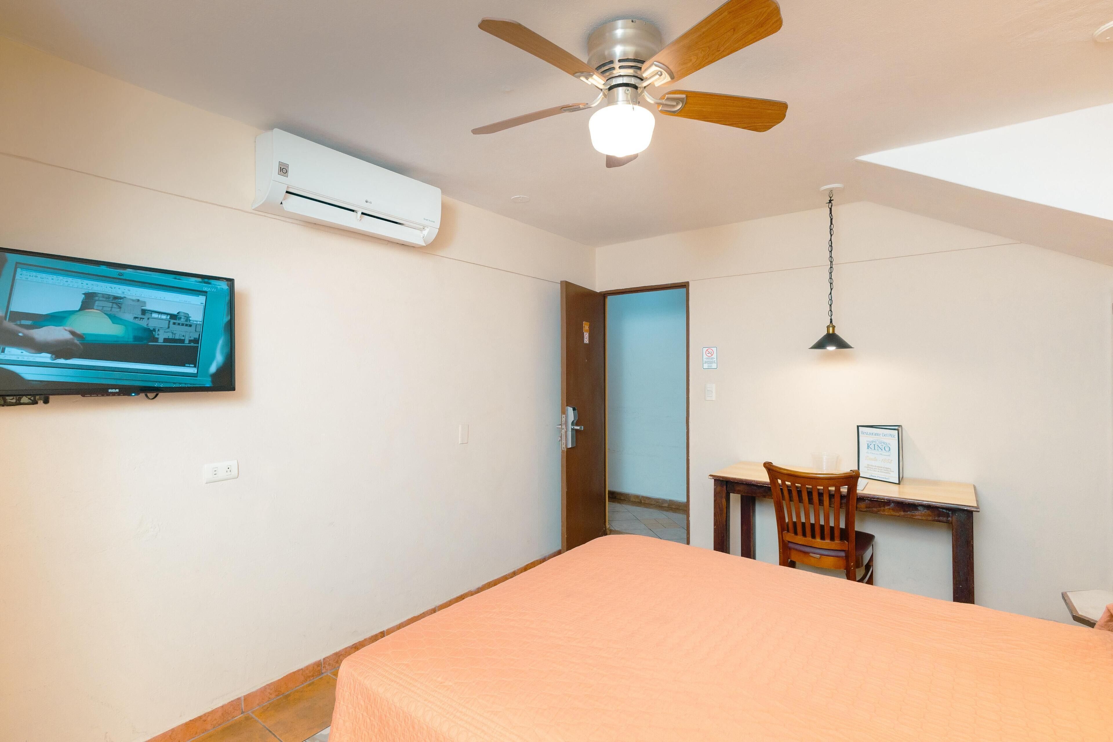 A bedroom with a bed, a desk, a chair, a ceiling fan, an air conditioner, and a TV mounted on the wall.