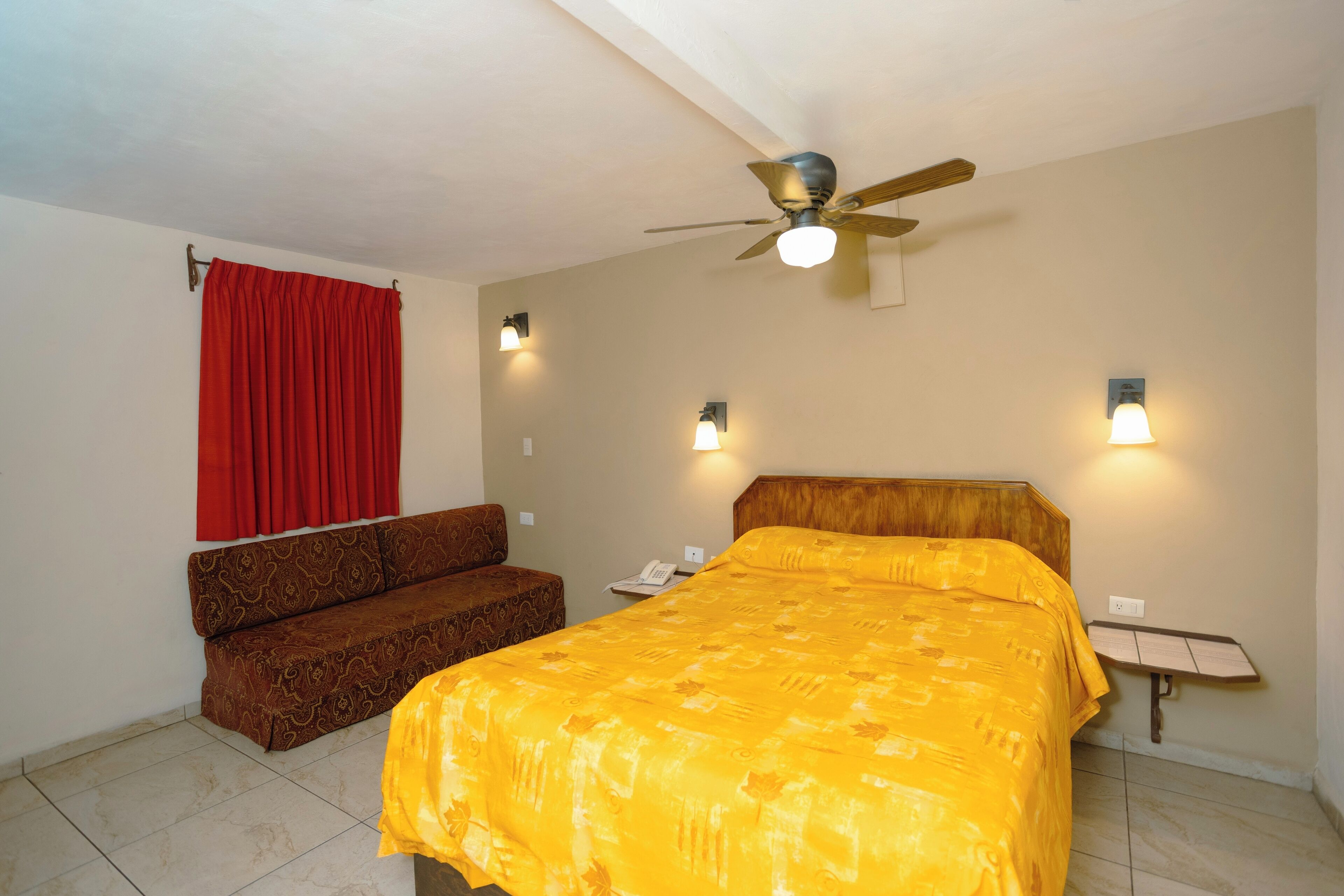 A hotel room with a bed, a sofa, a ceiling fan, and wall-mounted lights.