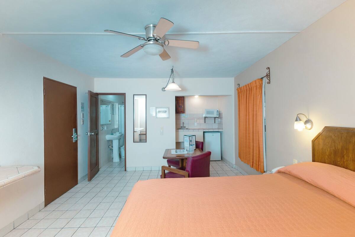 A hotel room with a bed, a desk with a chair, a kitchenette, a bathroom, and a ceiling fan.