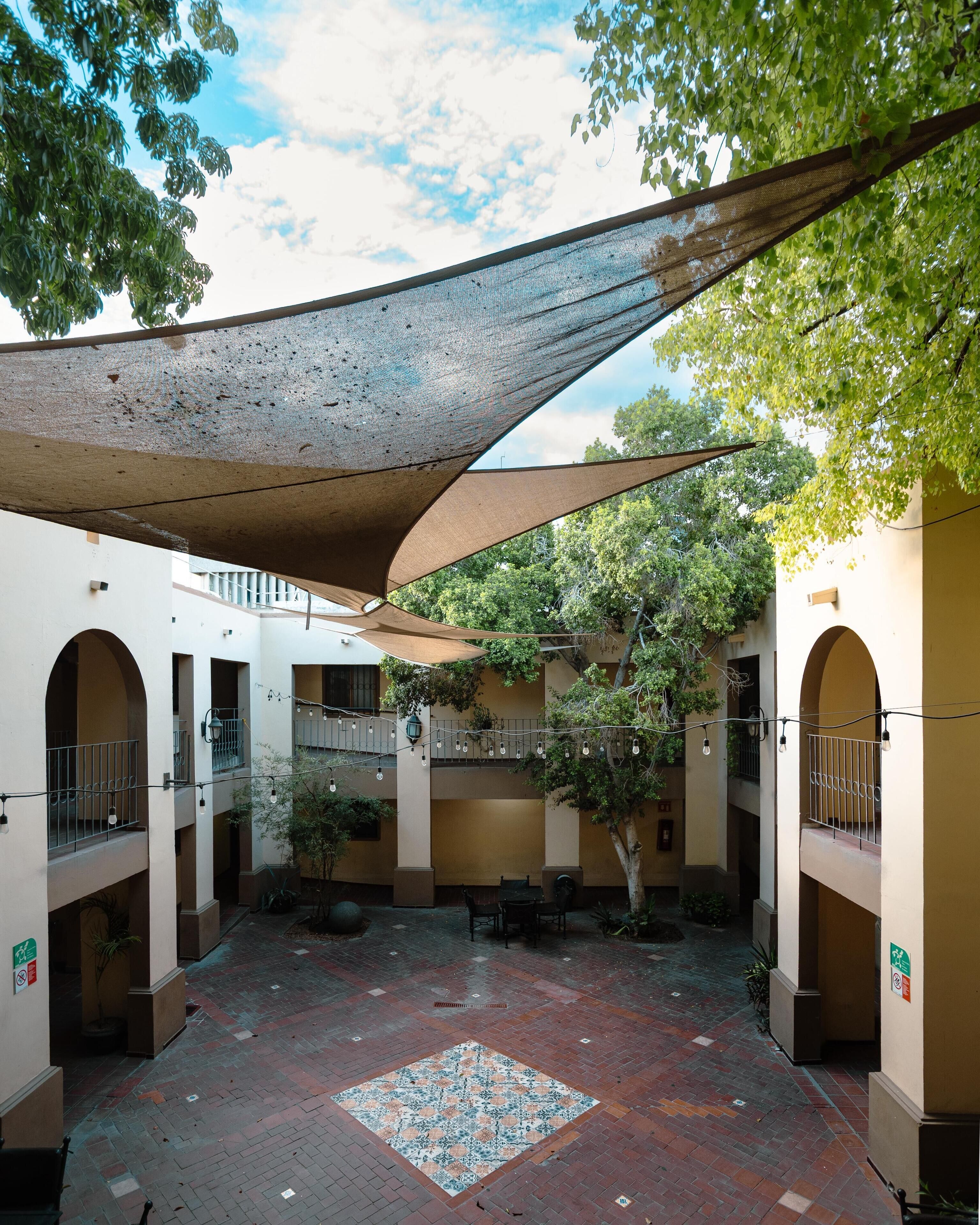 A courtyard with a patterned floor, shaded by a large fabric canopy, surrounded by walls with arched doorways and balconies.