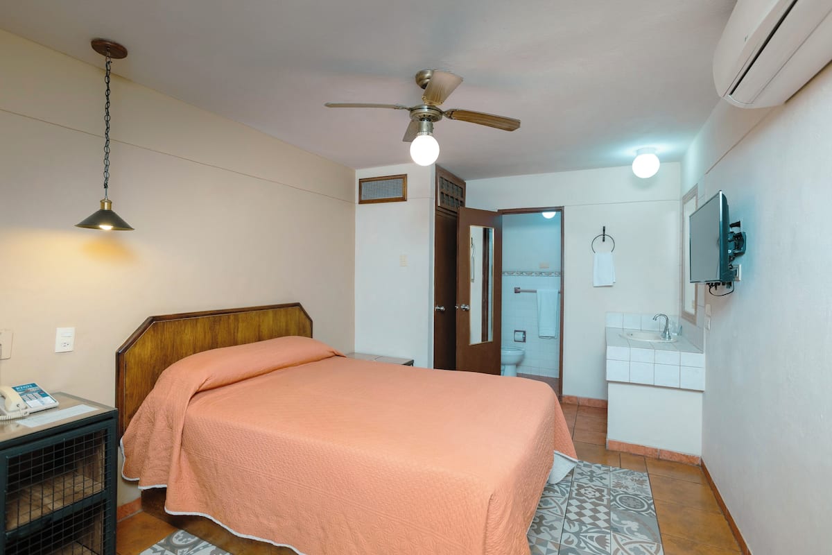 A bedroom with a bed, a ceiling fan, a bedside lamp, a desk with a phone, and a bathroom with a sink and mirror.