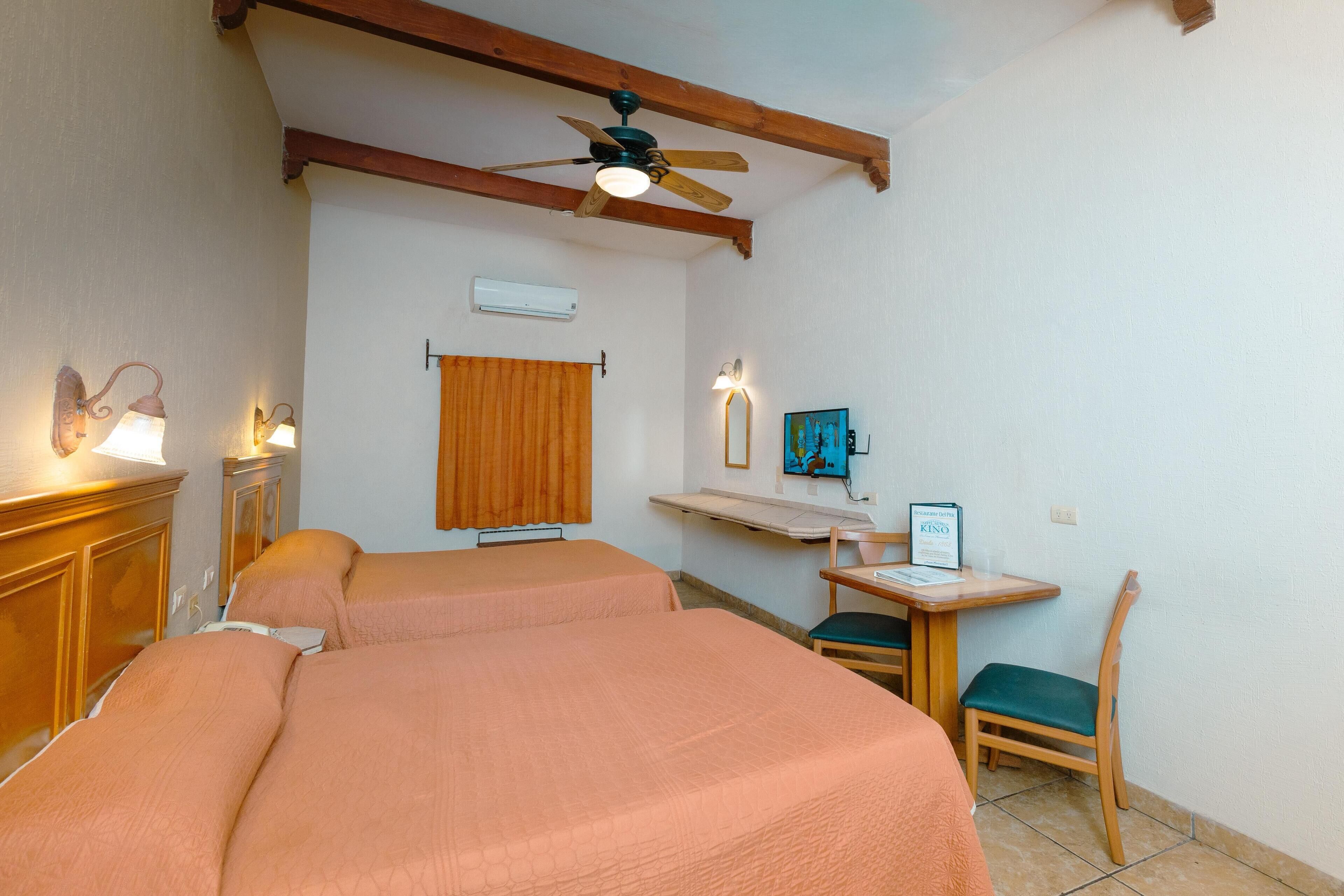 Habitación de hotel con dos camas, un ventilador de techo, una mesita con una silla y un estante de pared con un marco de foto y un cartel.