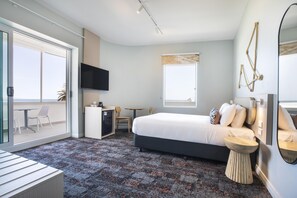 Una habitación de hotel moderna con una cama grande, un escritorio, un televisor y un balcón con mobiliario exterior.
