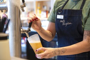 A person wearing a name tag and an apron, holding a glass of beer near a tap. - Nightcap at Esplanade Hotel (Brighton)