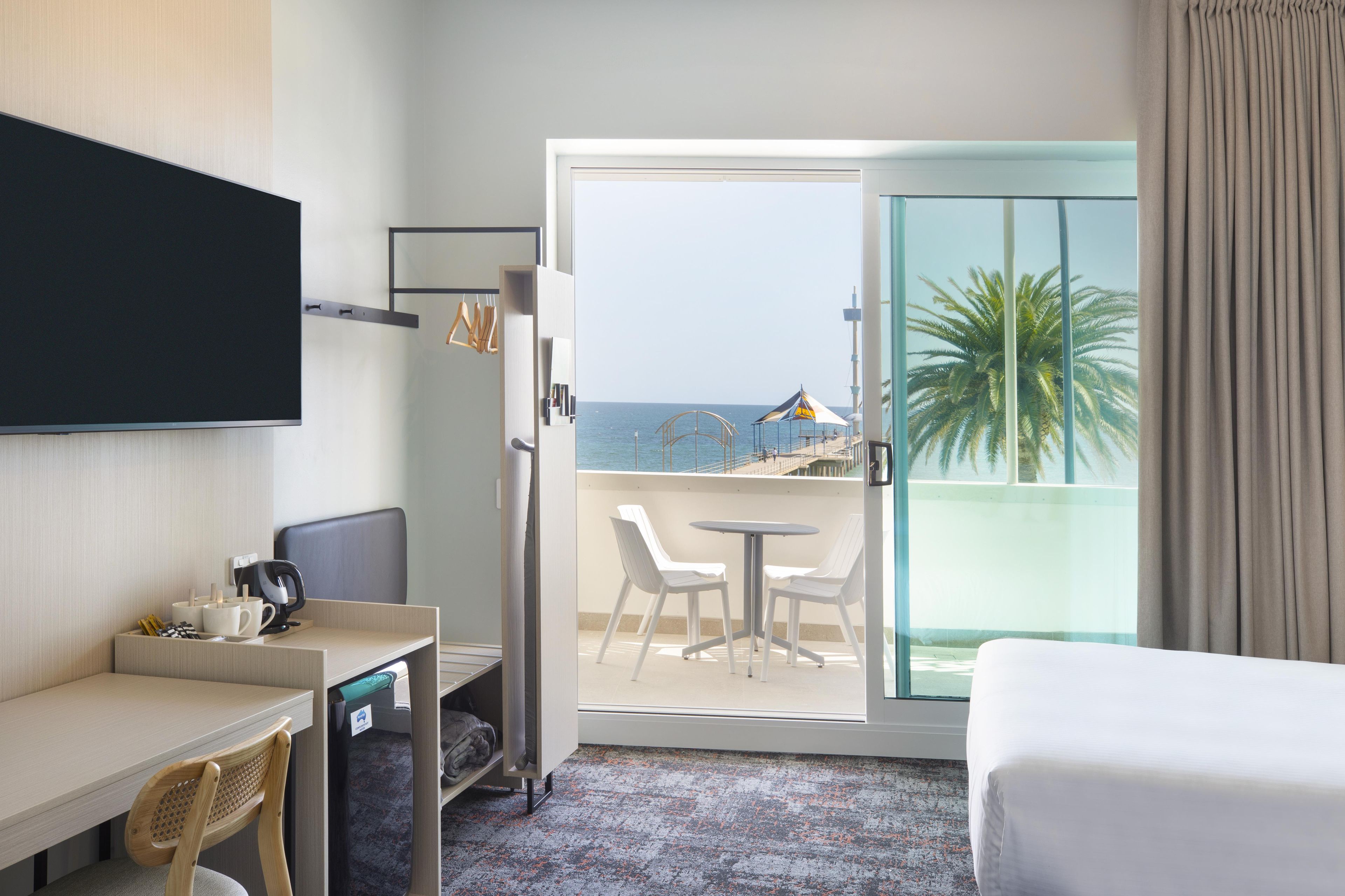 Habitación de hotel con cama, escritorio con silla, televisor montado en la pared, balcón con vista al mar y palmeras, y puerta corredera de vidrio.