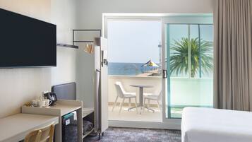 Habitación de hotel con cama, escritorio con silla, televisor montado en la pared, balcón con vista al mar y palmeras, y puerta corredera de vidrio.