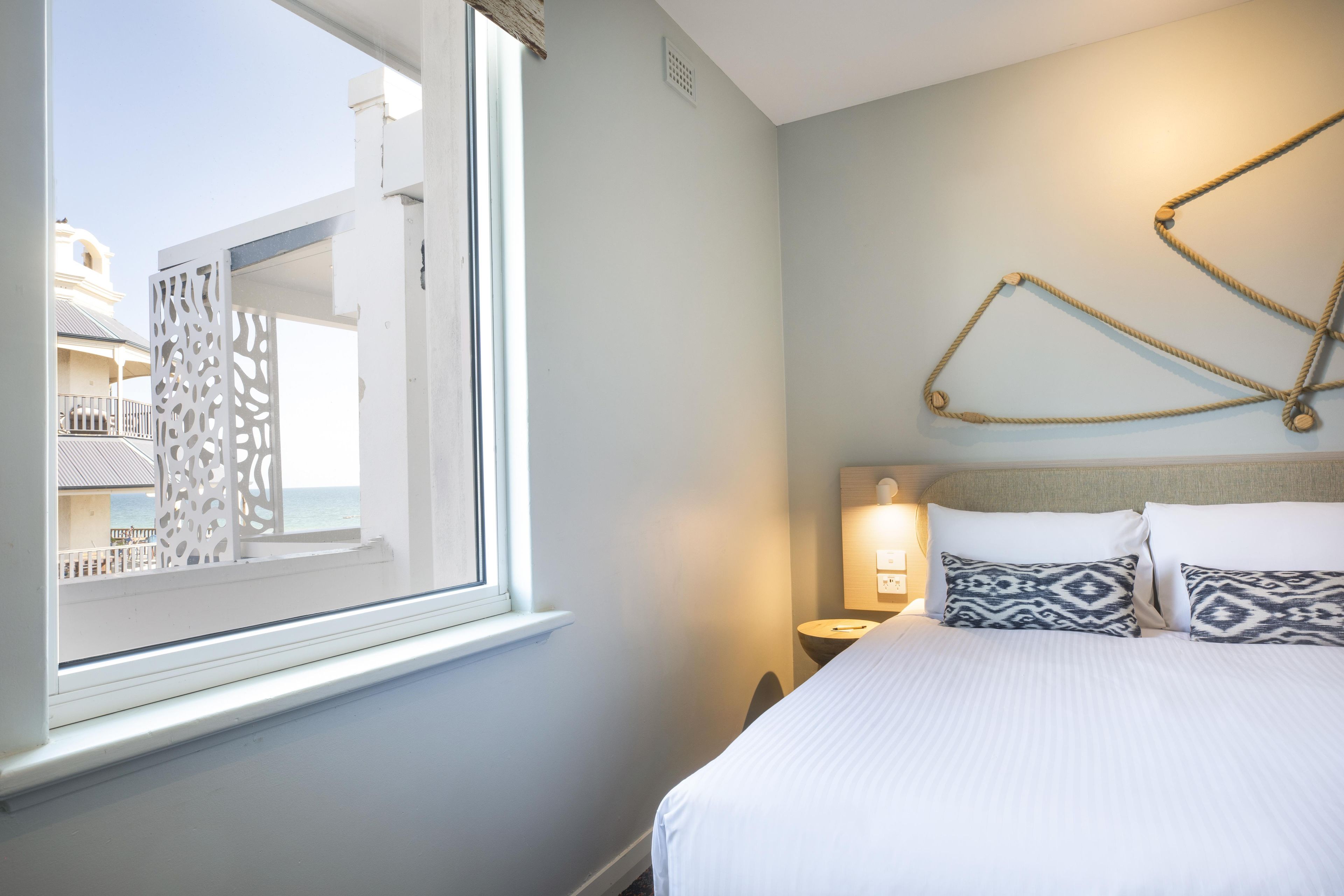 Habitación de hotel con cama, mesita de noche, lámpara de pared y vista a un balcón con barandilla.
