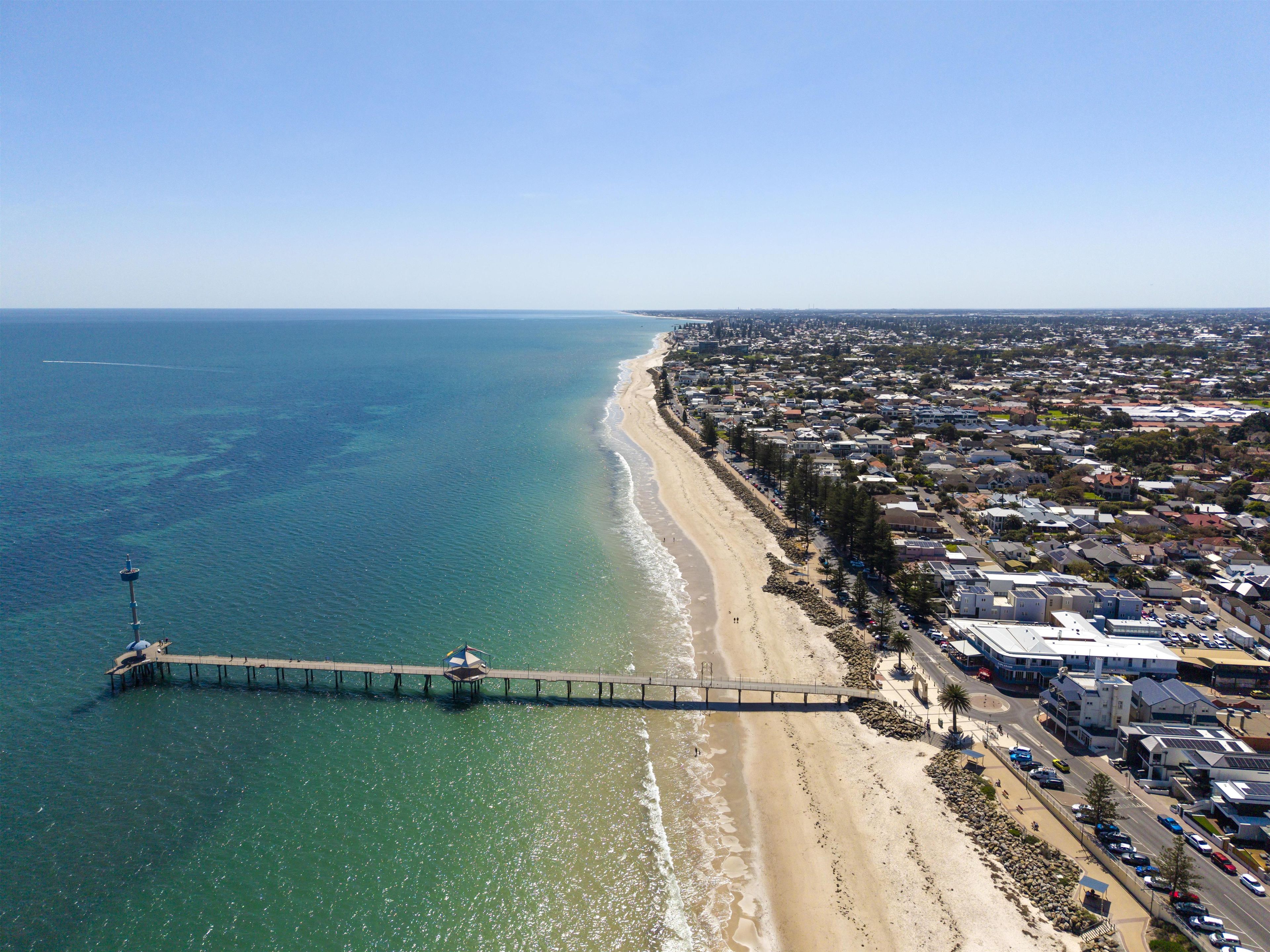 An aerial view of a coastal area with a pier extending into the sea, adjacent to a beach and residential buildings.