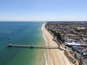 Vista aérea de una zona costera con un muelle que se adentra en el mar, junto a una playa y edificios residenciales.