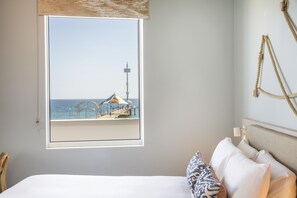 Un dormitorio con un ventanal que da a una playa, una cama con ropa blanca y vistas a un muelle con una pequeña estructura.