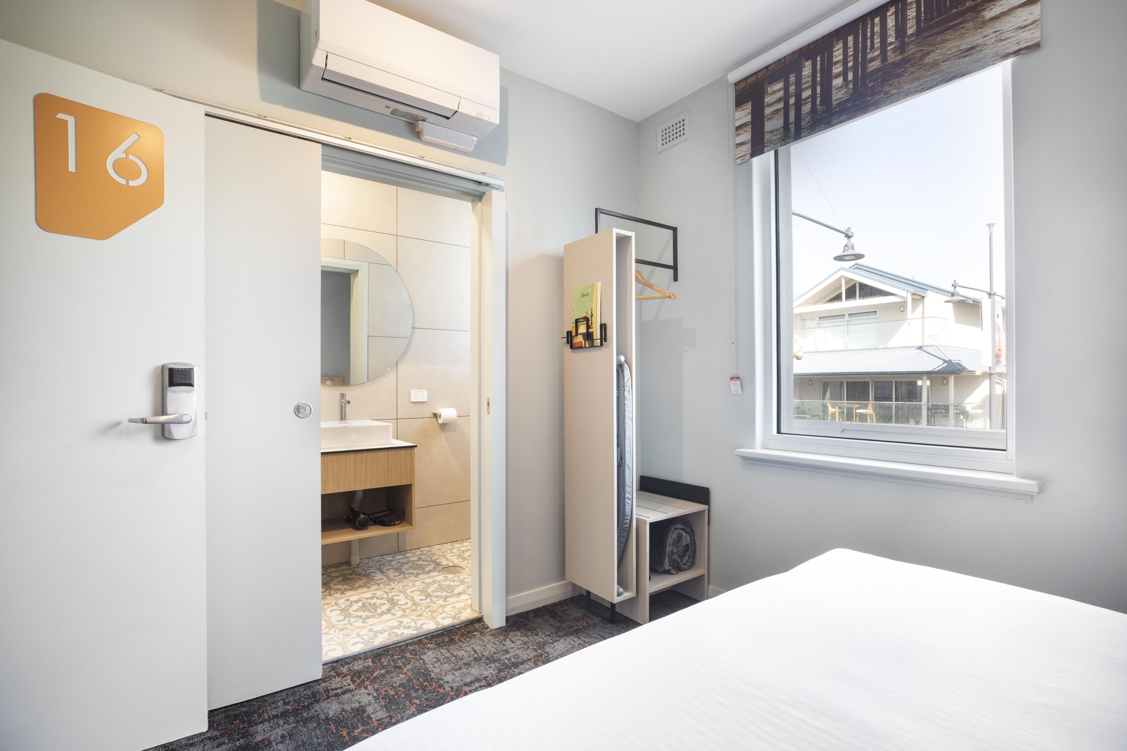 A hotel room with a bed, a bathroom with a toilet and sink, a mirror, and a window with a view of buildings.