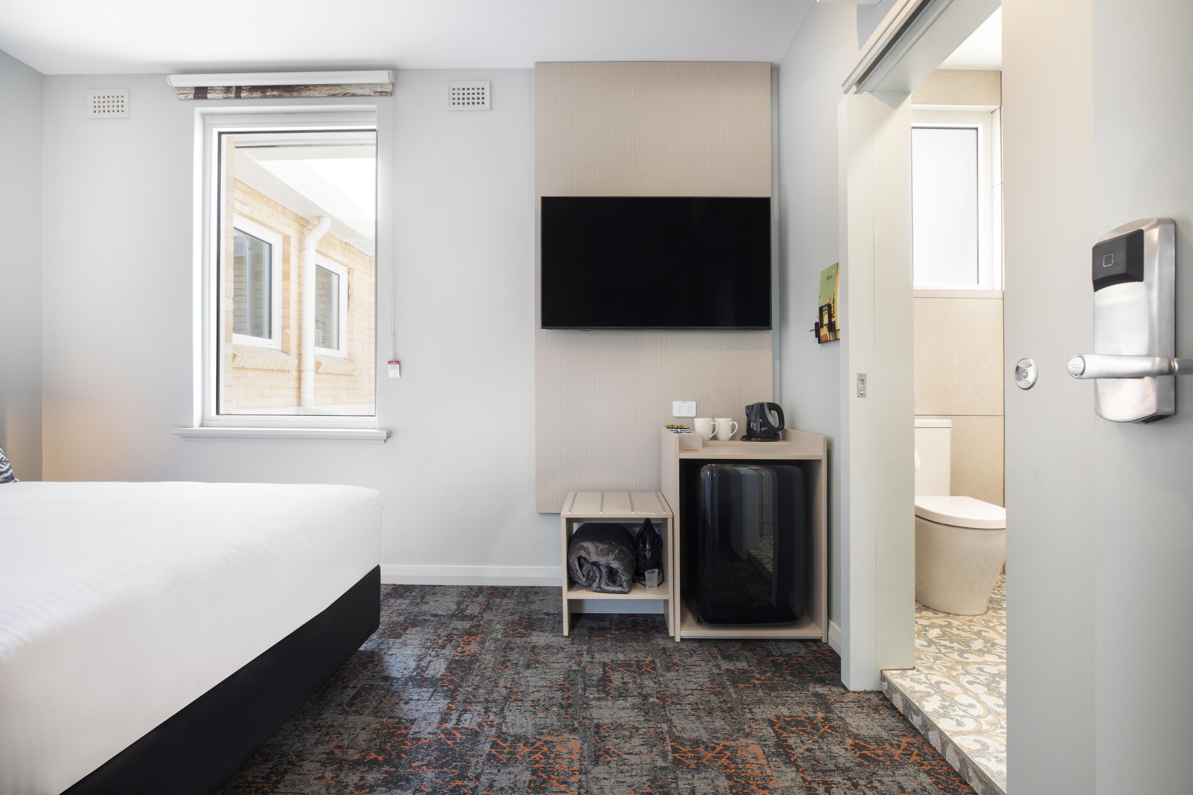 Habitación de hotel con cama, televisor de pantalla plana montado en la pared, una pequeña zona de estar con mesa y sillas, y baño con inodoro y lavabo.