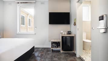 A hotel room with a bed, a flat-screen TV mounted on the wall, a small seating area with a table and chairs, and a bathroom with a toilet and sink.