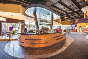 A modern bar area with a curved counter, multiple shelves of bottles, and a seating area with chairs and tables. - Nightcap at Sandown Park Hotel (Noble Park)